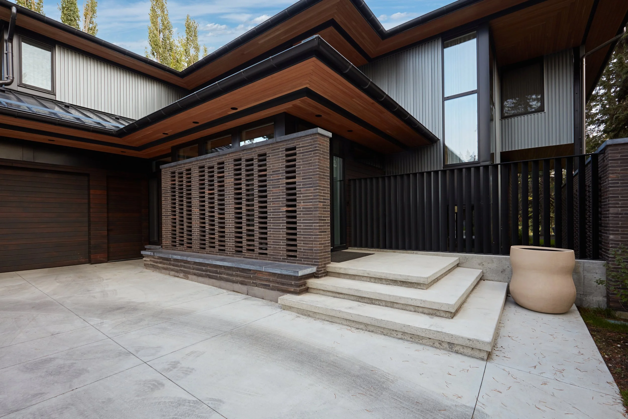 Modern house exterior with brick, metal, and wood elements, concrete steps, and surrounding trees.