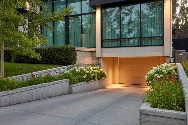 Modern house exterior with a driveway leading to a garage, flanked by concrete walls and landscaped with green bushes and white flowers.