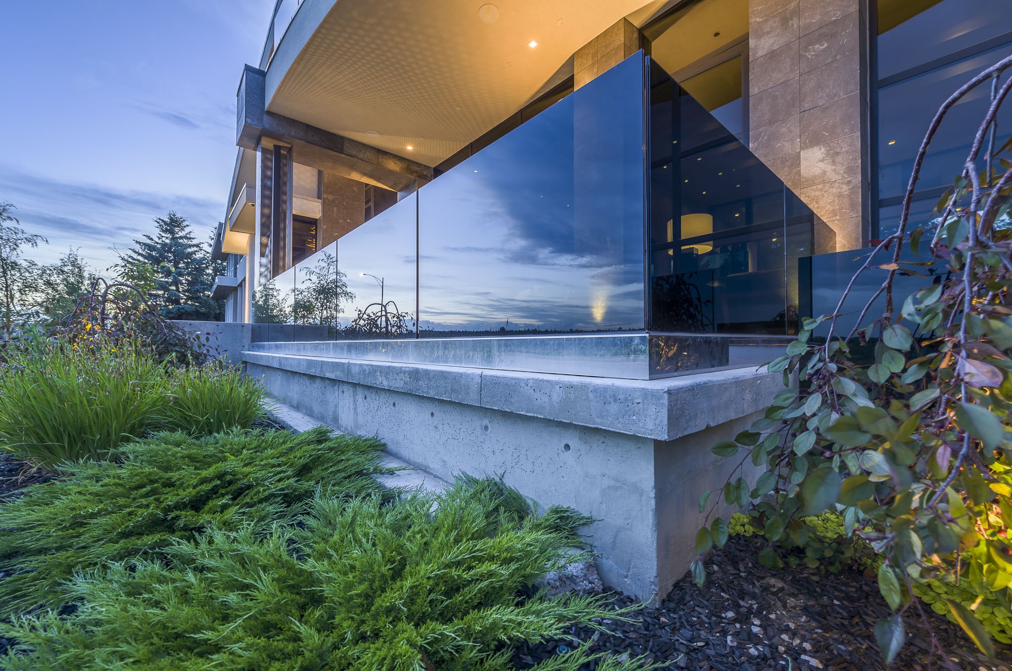 Modern building with large reflective glass windows and a concrete foundation, surrounded by lush green bushes and plants, under a cloudy evening sky.