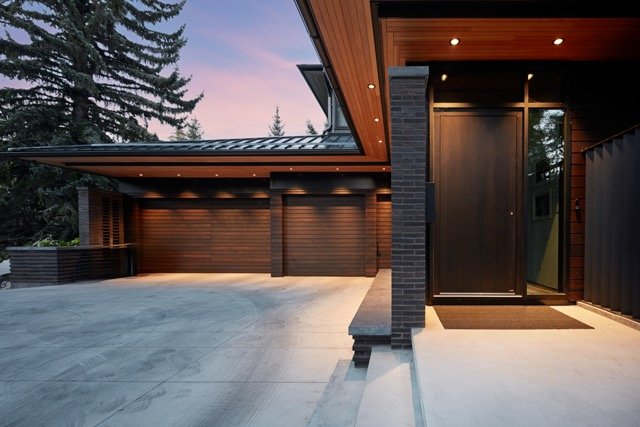 Modern house exterior with tall trees, featuring a wooden facade, large entry door, and a spacious driveway at sunset.