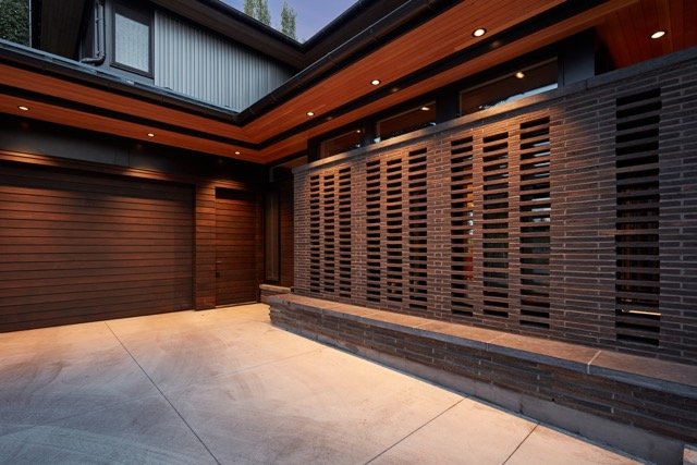 Modern house exterior with wooden paneling and brick lattice wall, featuring recessed lighting and concrete driveway.