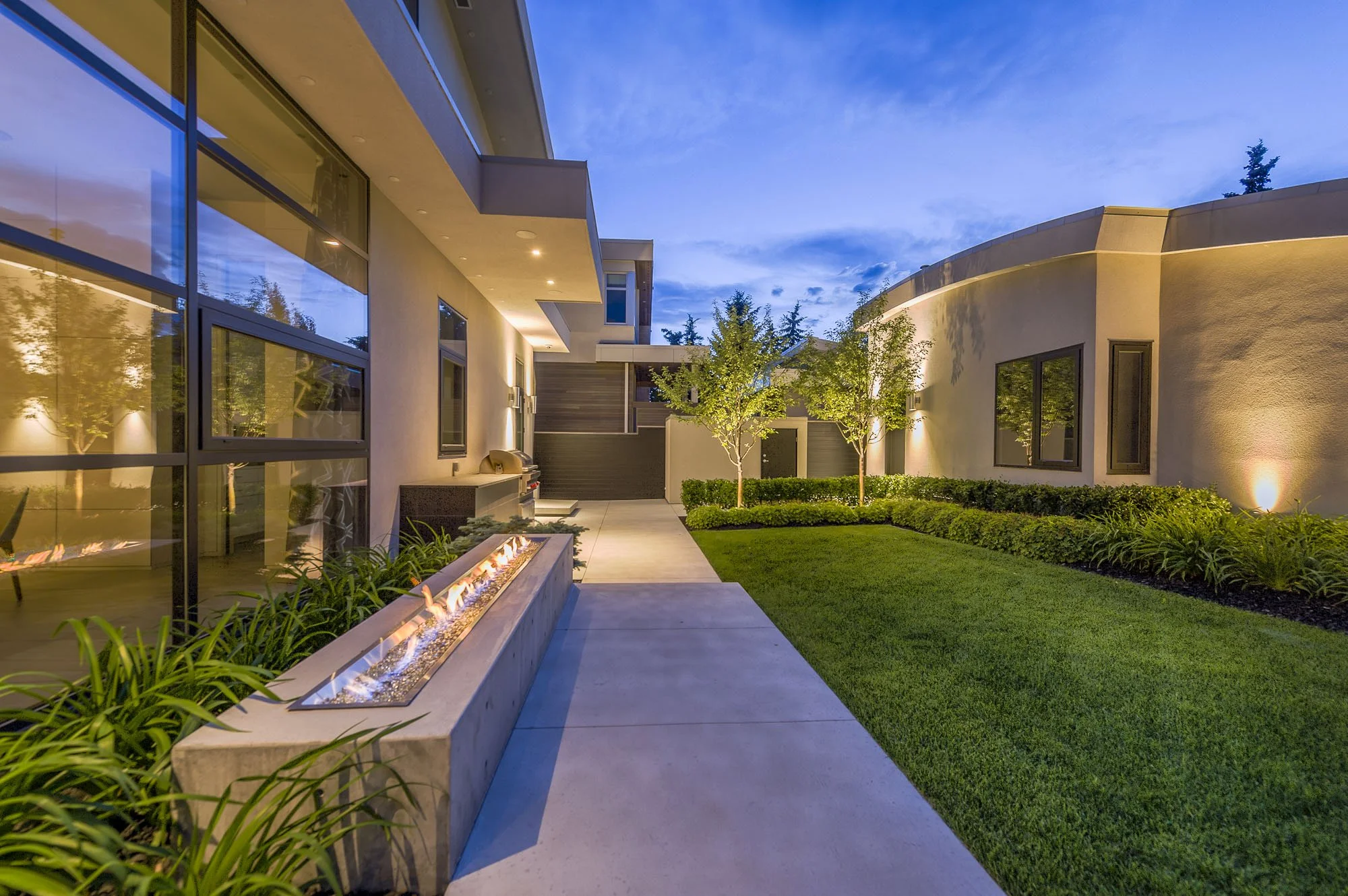Modern residential backyard at dusk with lit landscaping, two small trees, and a fire feature along a paved walkway, adjacent to a contemporary house with large windows and exterior lighting.
