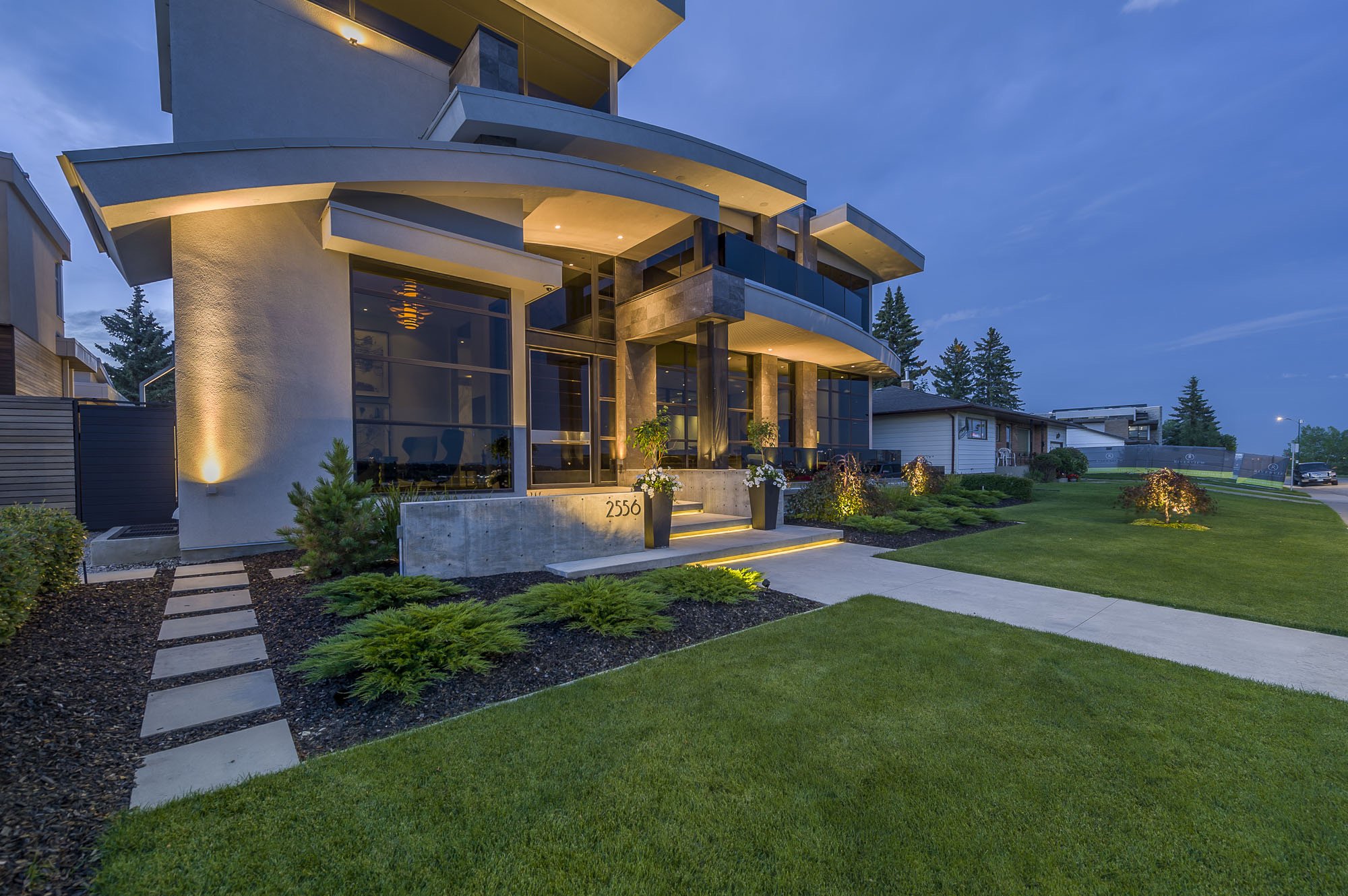 Modern multi-story house with large glass windows and balconies, surrounded by a well-maintained lawn and garden, during dusk with outdoor lighting.