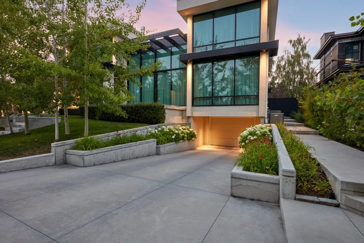Modern residential building with large glass windows, concrete landscaping, and an underground garage entrance, surrounded by greenery and flowerbeds.