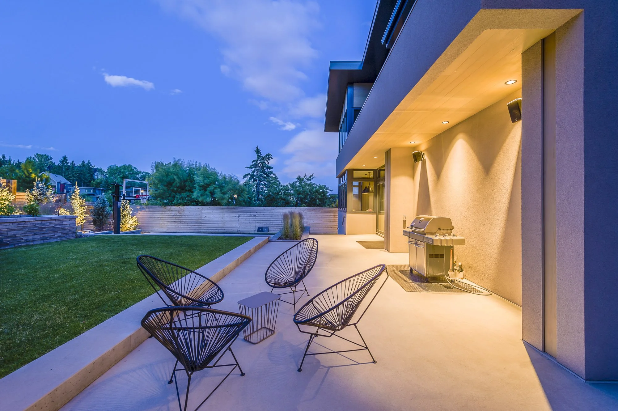 Modern house patio with outdoor seating, a barbecue grill, and a well-maintained backyard with a basketball hoop, trees, and a wooden fence during evening with exterior lighting.