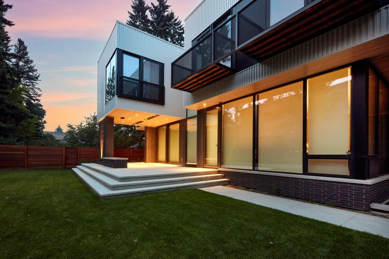 Modern two-story house with large glass windows, a small outdoor staircase leading to a patio, and a well-maintained lawn at sunset.