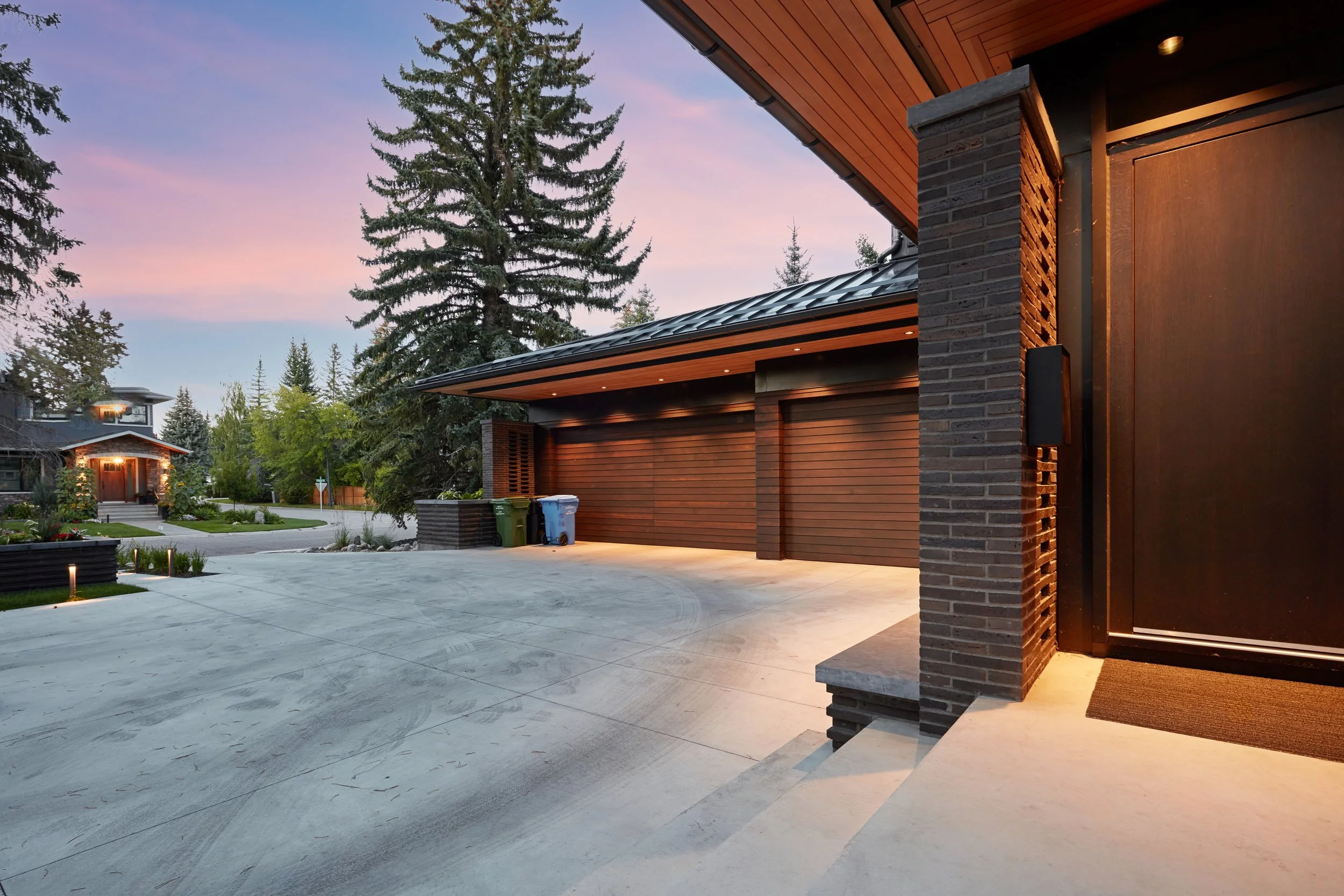 Modern house with a brick pizza oven on the porch, surrounded by trees under a purple sky at sunset, with trash bins outside and a manicured yard in the background.