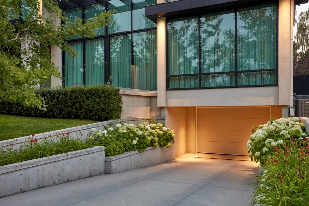 Modern house with large glass windows, landscaped garden with bushes and flowers, and a garage on the lower level illuminated by warm lighting.