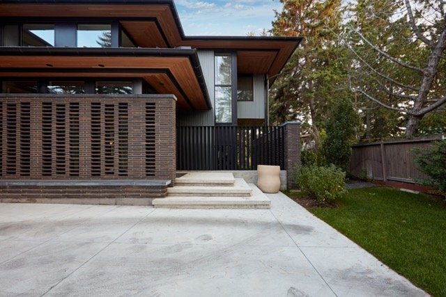 Modern house exterior with brick and wood elements, featuring a unique brick lattice wall, concrete steps leading to the entrance, a large ceramic planter, and landscaped garden with grass and trees.