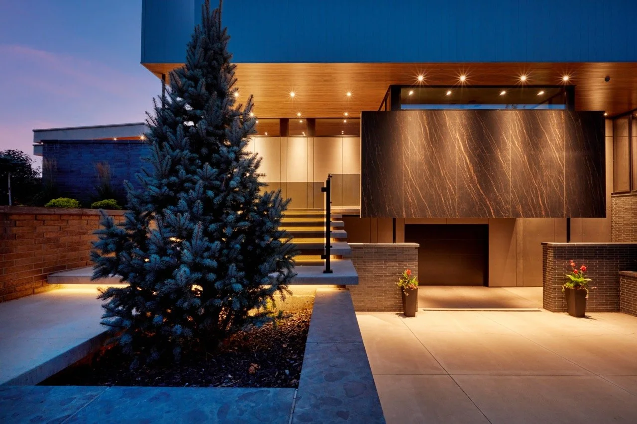 Modern house exterior at dusk with a large blue spruce tree, lit stairs, and a marble accent wall.