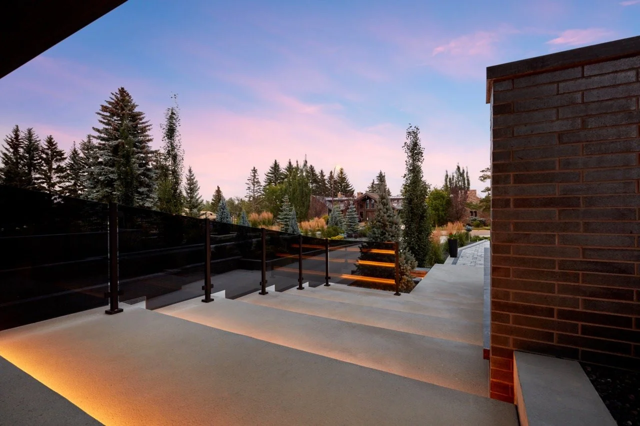 View from a modern house balcony at sunset, showing a landscaped yard with tall trees and houses in the background, illuminated stairs, and a brick wall.