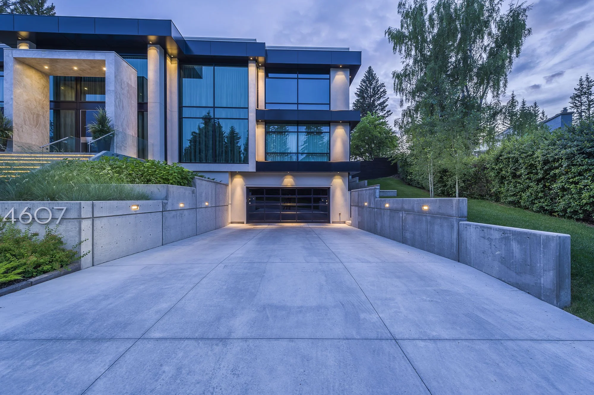 Modern residential house with large glass windows, concrete driveway, landscaping, and trees surrounding property, during twilight.