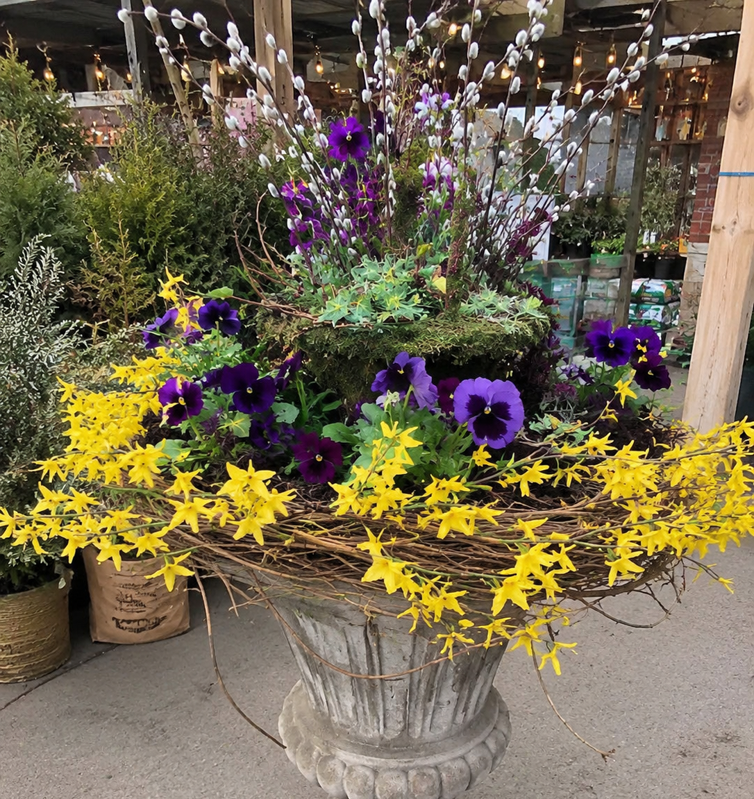 Vase filled with purple, yellow, and white flowers, including violets and pussy willows, arranged in a decorative outdoor setting.