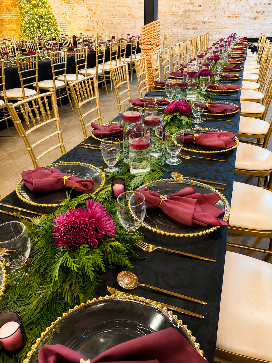 A long banquet table set for a festive holiday celebration. The table has a black tablecloth, with gold chargers, gold flatware, white napkins, and glassware. A lush green garland with pinecones and white candles runs down the center. In the background, there are gold chairs and a brick wall with holiday decorations including black, white, and yellow balloons.