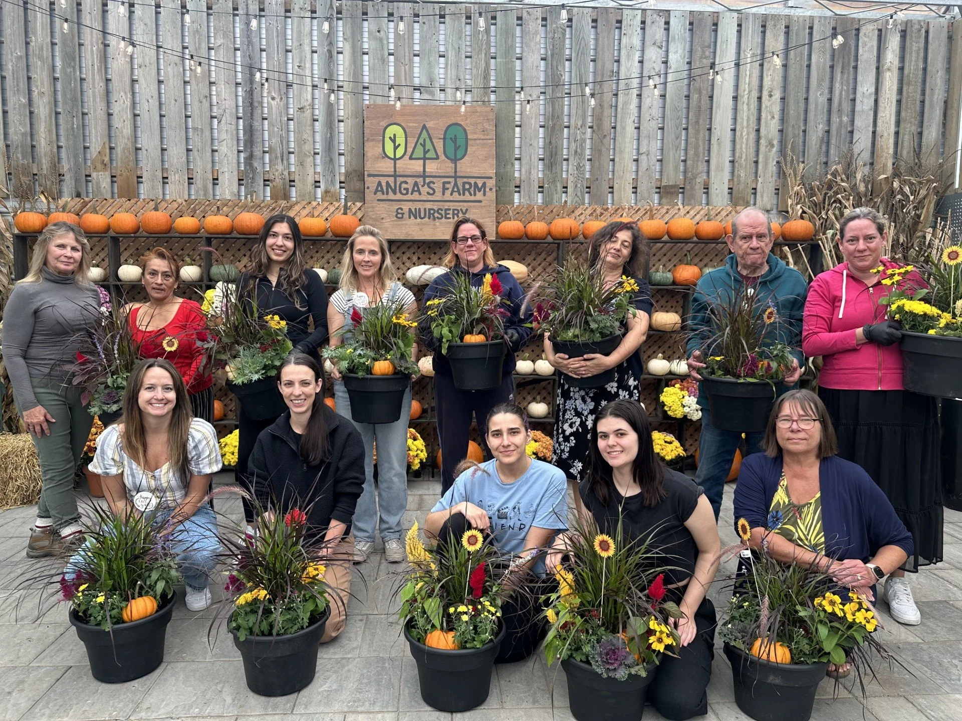 A group of people sitting at a long table in a greenhouse for an event at Anga's Farm & Nursery, with pink and white flowers and greenery decorating the space.