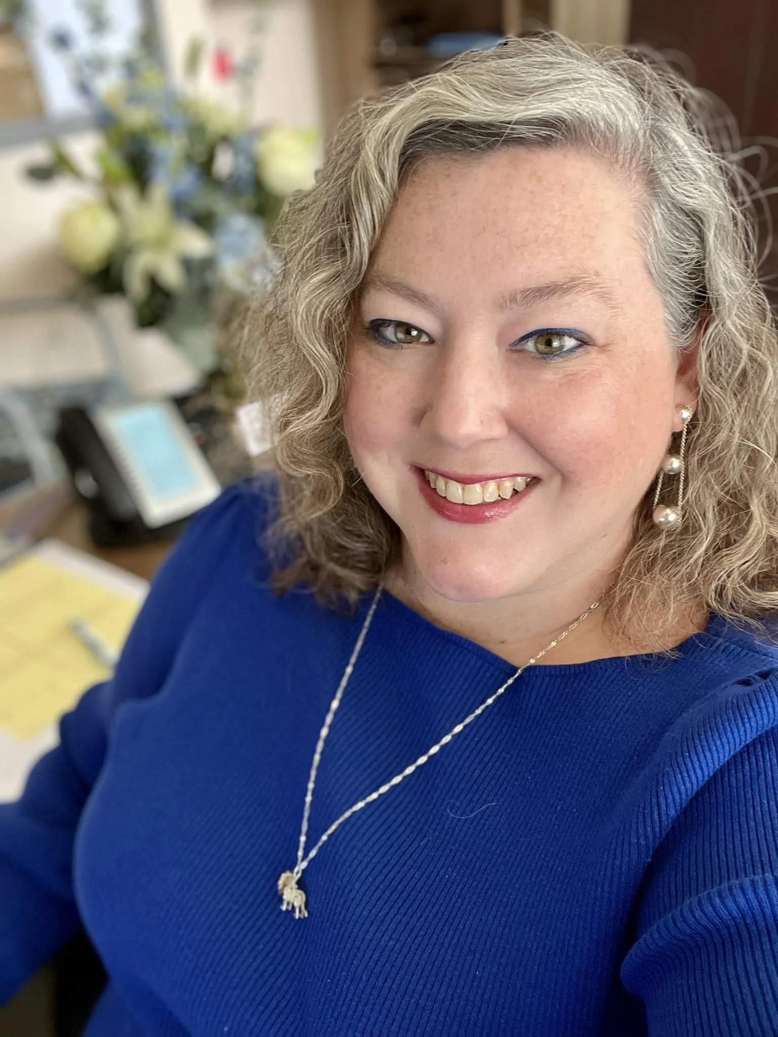 Selfie of a white woman with curly, shoulder-length hair that is a mix of light brown and gray. She is smiling, wearing a bright blue sweater and a silver necklace with a pendant. She also has pearl earrings. There is a blurred background.