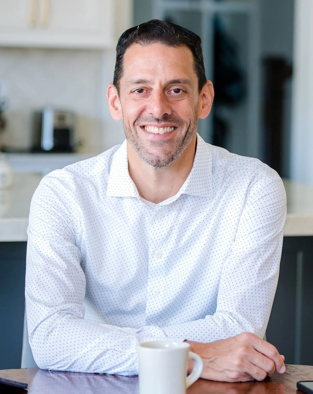 Headshot of a man sitting at a table. He has short dark hair and a light beard, and he is smiling at the camera. He is wearing a white shirt with small blue polka dots. There is a white mug in front of him on the table. The background is a kitchen.