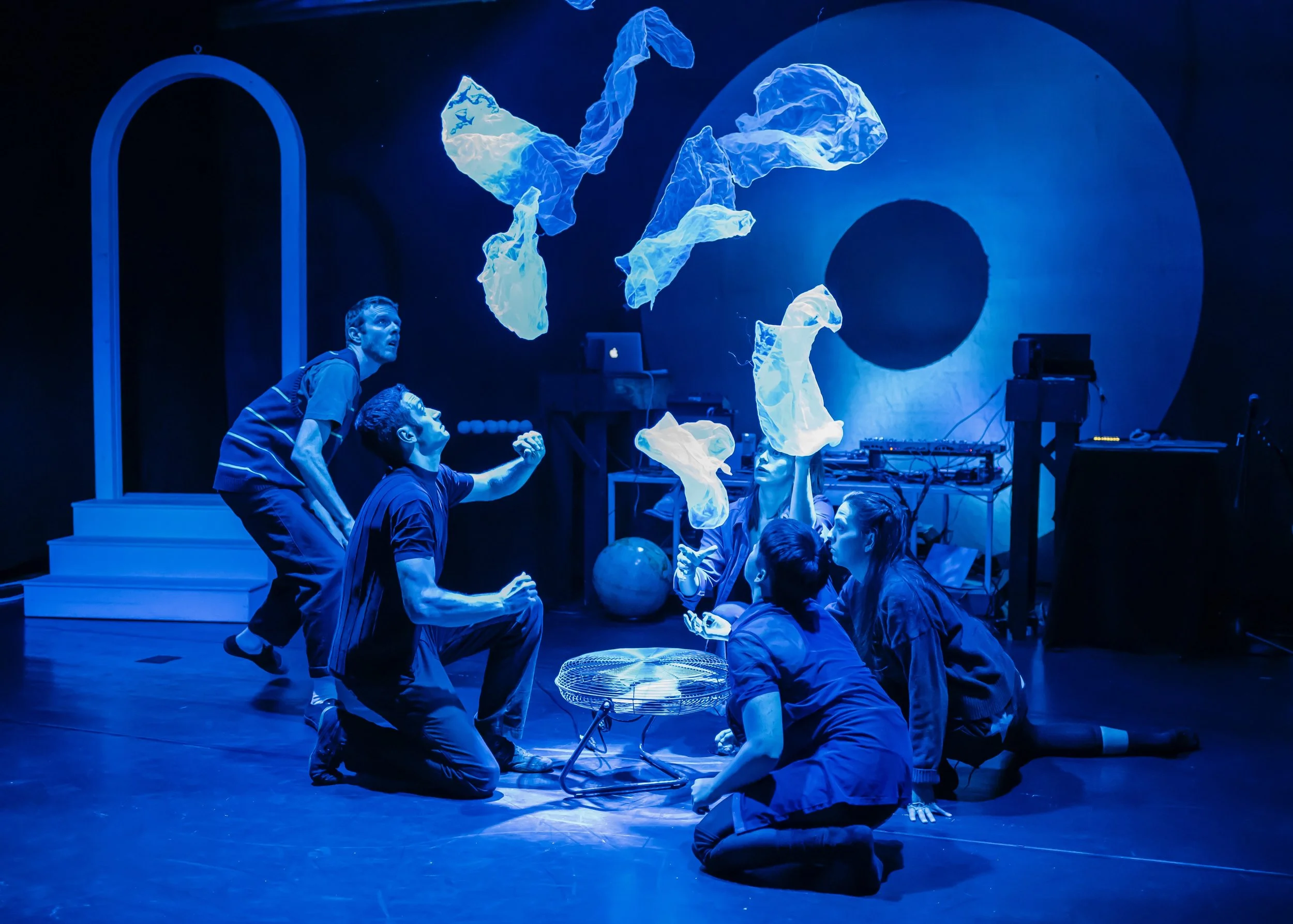 A theatrical performance with five actors on stage under blue lighting. Some actors are kneeling or sitting, looking up at hanging fabric pieces illuminated in the air. The background includes steps, a large circular projection, and electronic equipm