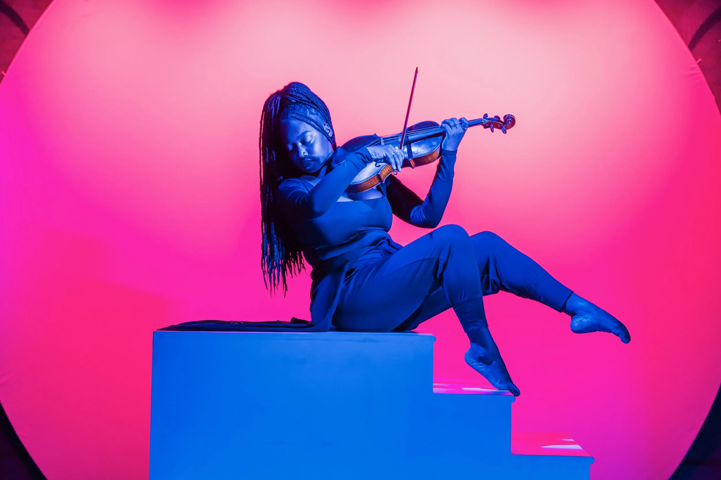 A woman with long braided hair playing the violin, sitting on a white step with a pink background