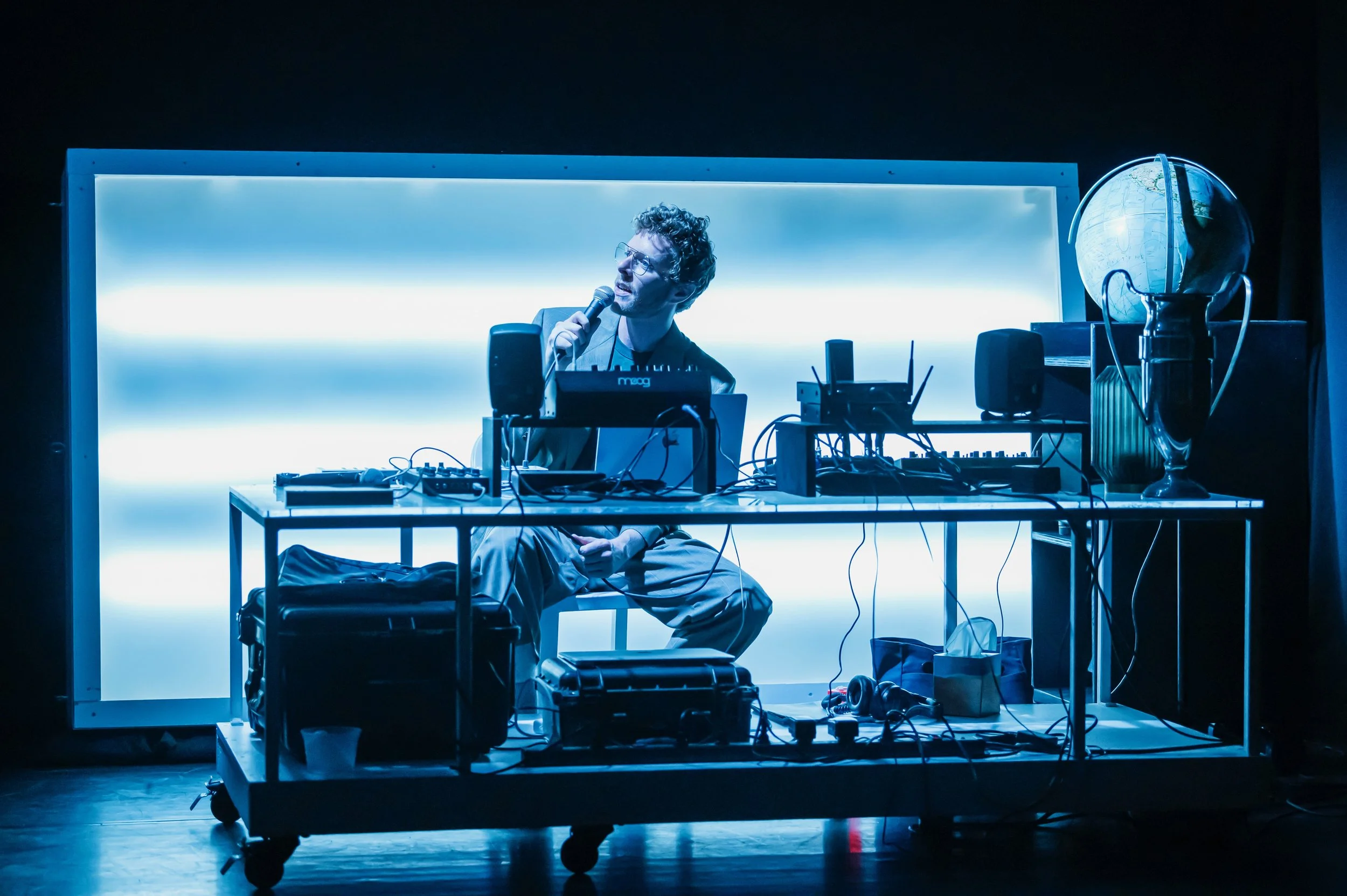 A man sitting on a chair behind a DJ booth with electronic equipment, holding a microphone, with a glowing blue background, and a globe on a stand to the right.