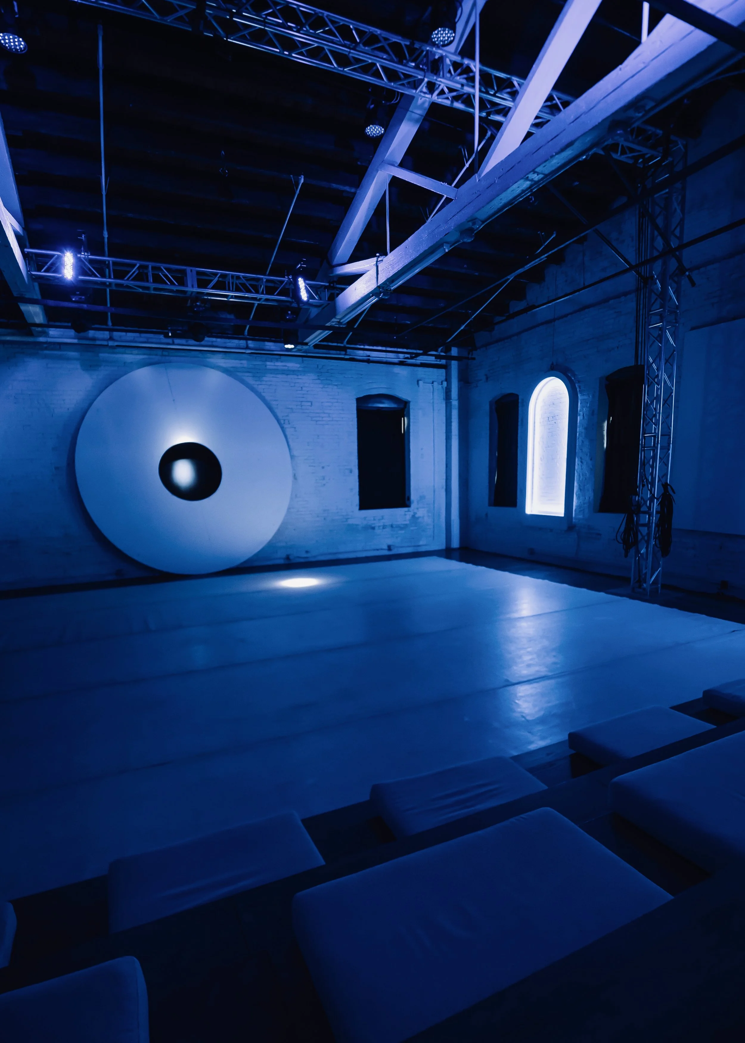An empty art gallery with blue lighting, featuring a large circular sculpture on the back wall, brick walls with windows, and padded benches on the floor.