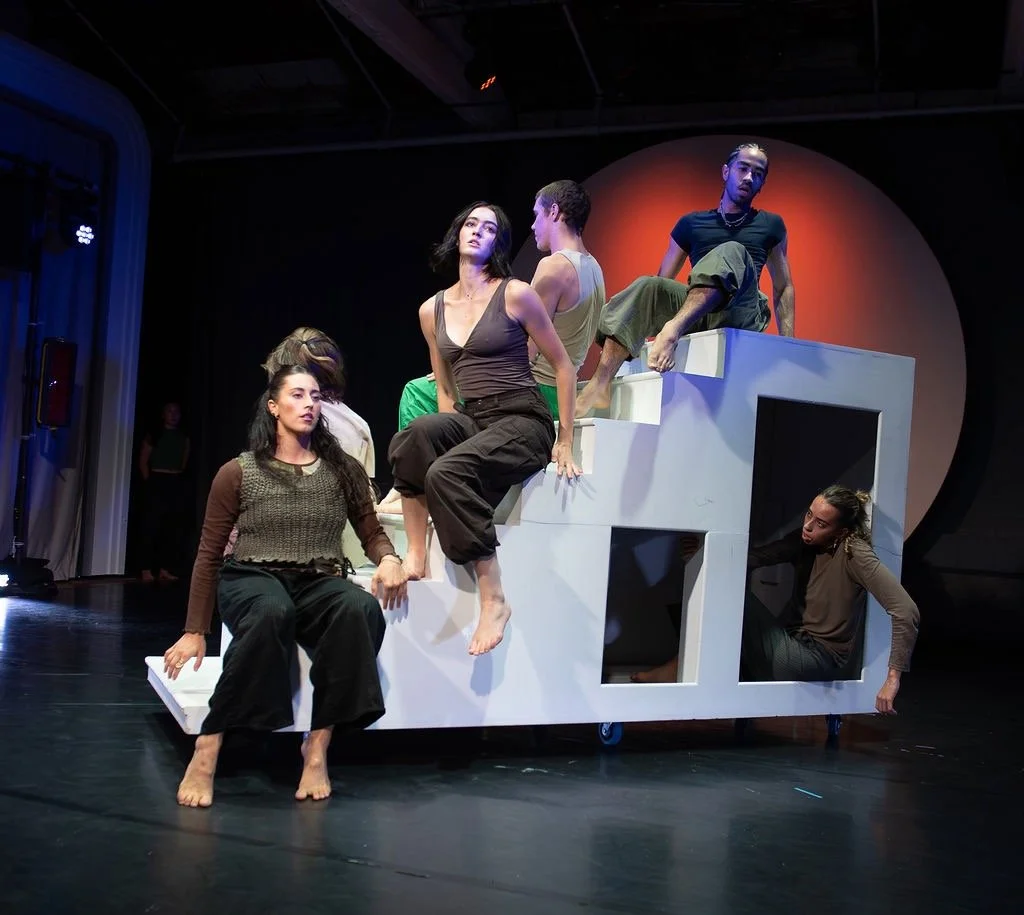 A group of seven people are on a stage with a black background, some sitting, standing, or climbing on a white, abstract structure with square and rectangular openings. The scene appears to be part of a theatrical performance or rehearsal.