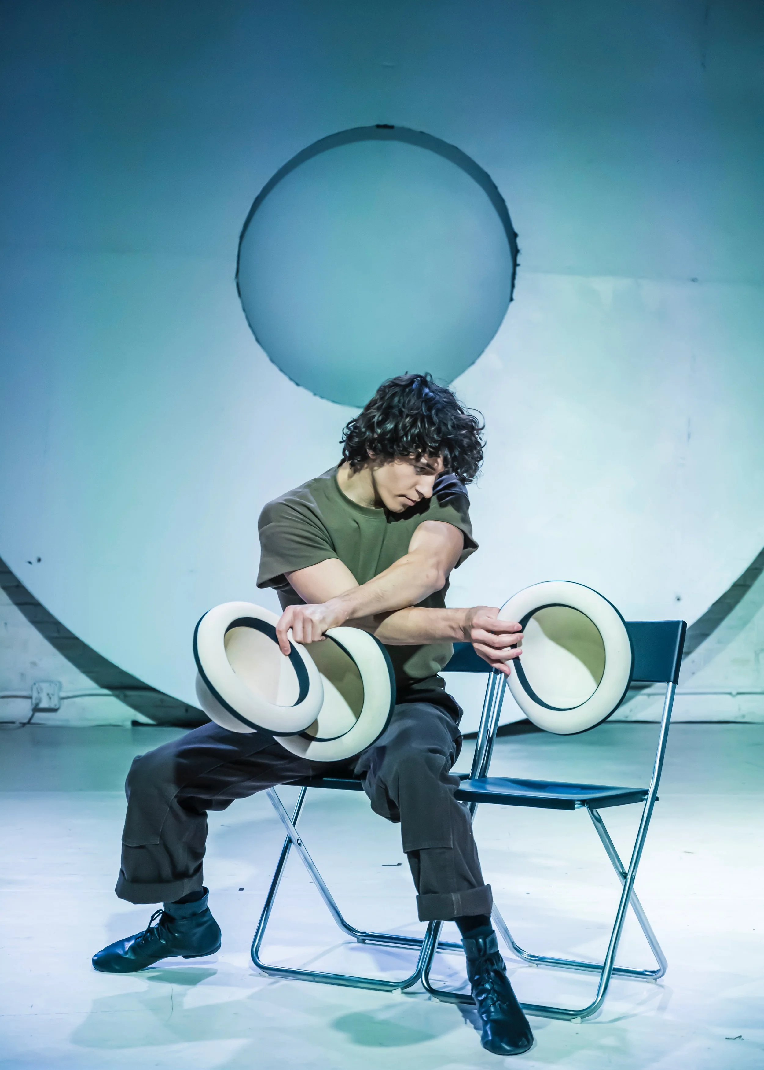 A person with curly hair sitting on a chair, holding white circular objects, with a large circular ceiling or backdrop behind them.