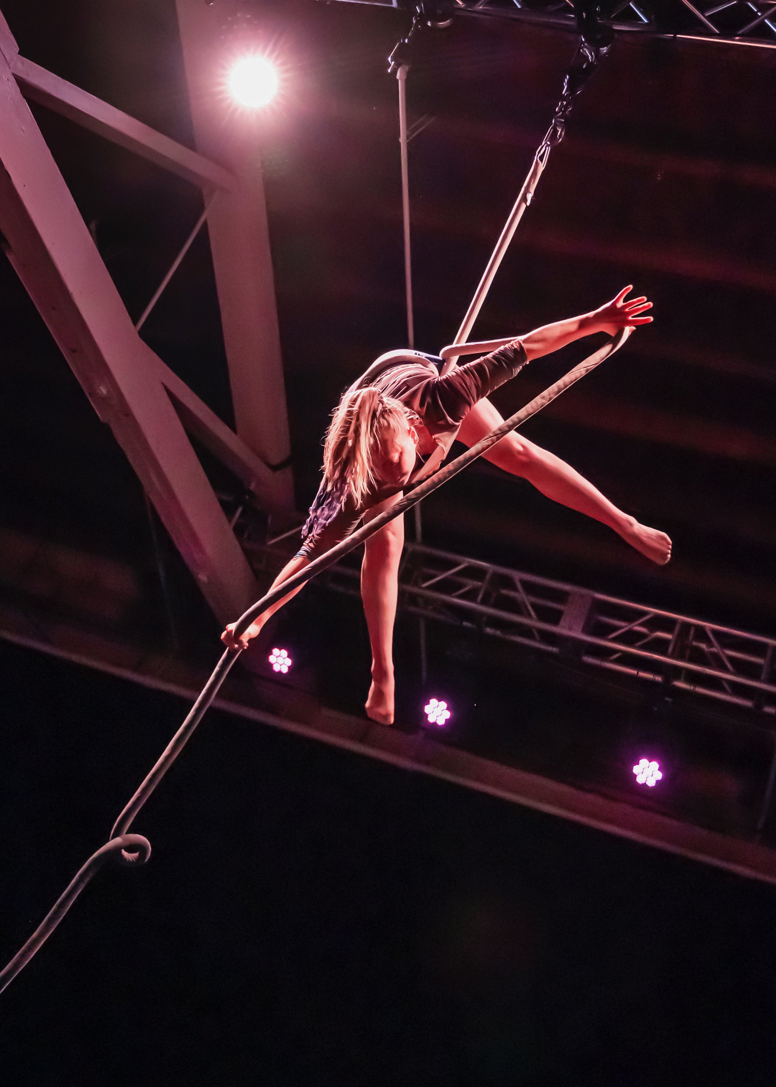 A female aerialist performing on a trapeze during a circus act with stage lights shining down.