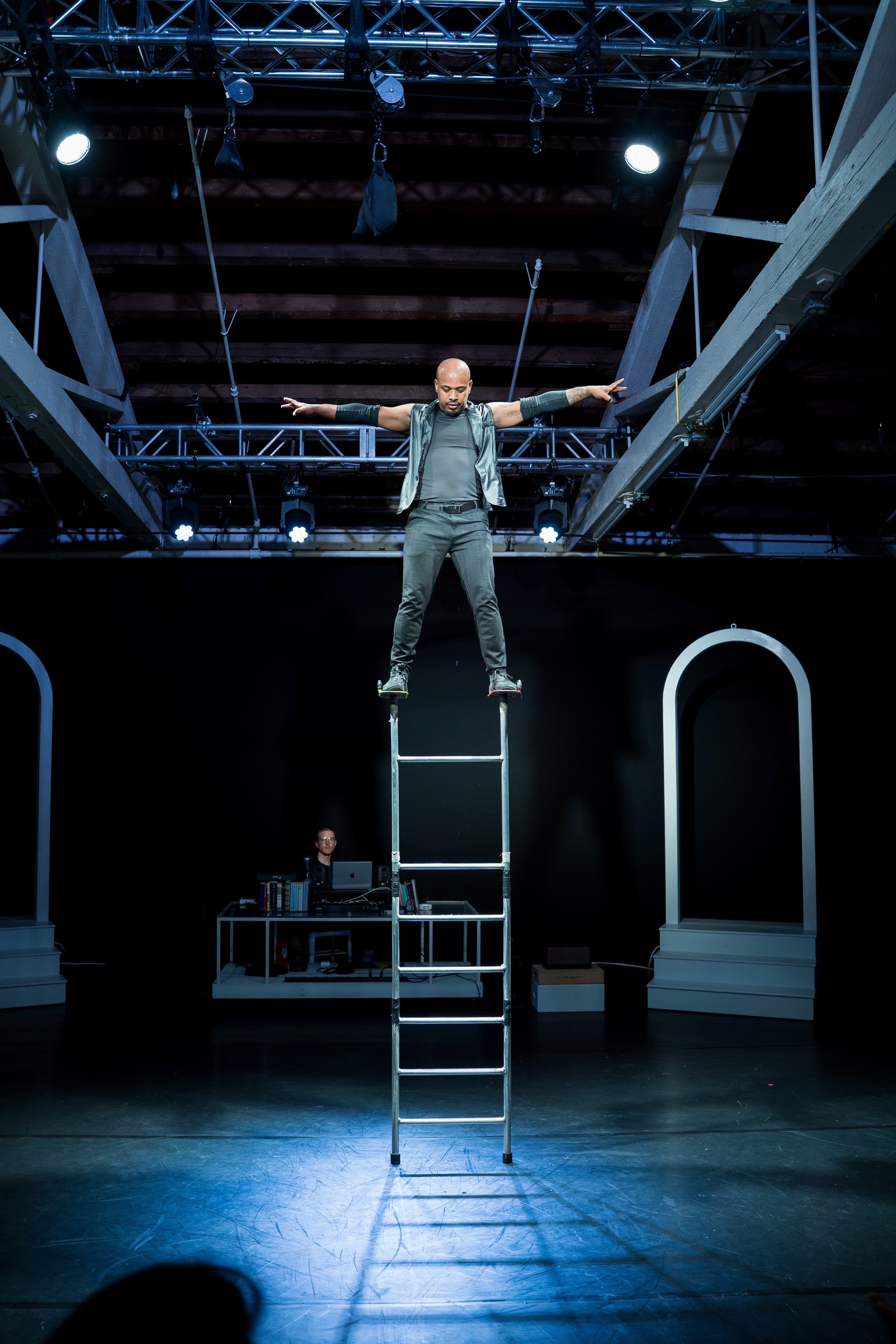 A performer balancing on a ladder on stage with arms outstretched and a stage setup in the background.