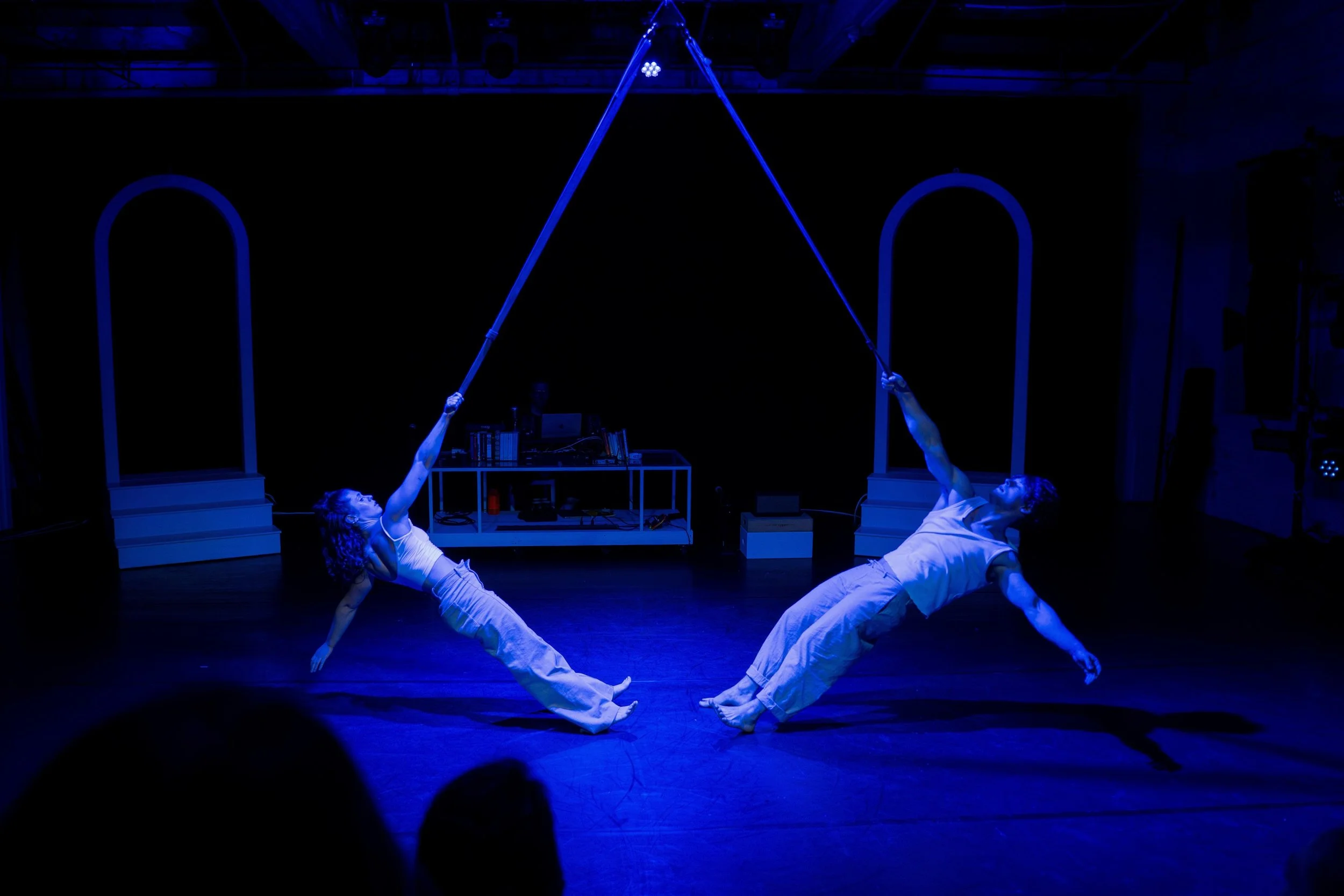 Two performers doing a trapeze act on stage in blue lighting, with their bodies extended and connected by cables, in a dance or acrobatic routine.