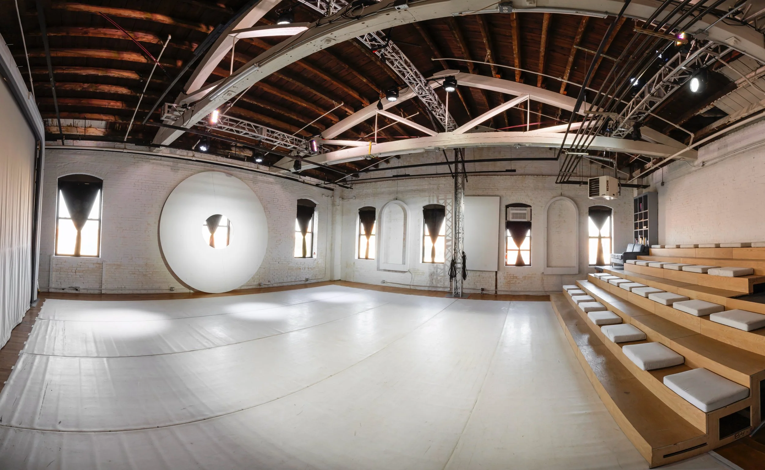 Empty event or performance space with a polished wood floor, white brick walls, and arched windows with black curtains. A large circular art piece with a glowing center is on one wall, and there are tiered seating with white cushions along one side. The ceiling has exposed wooden beams, lighting, and technical equipment.