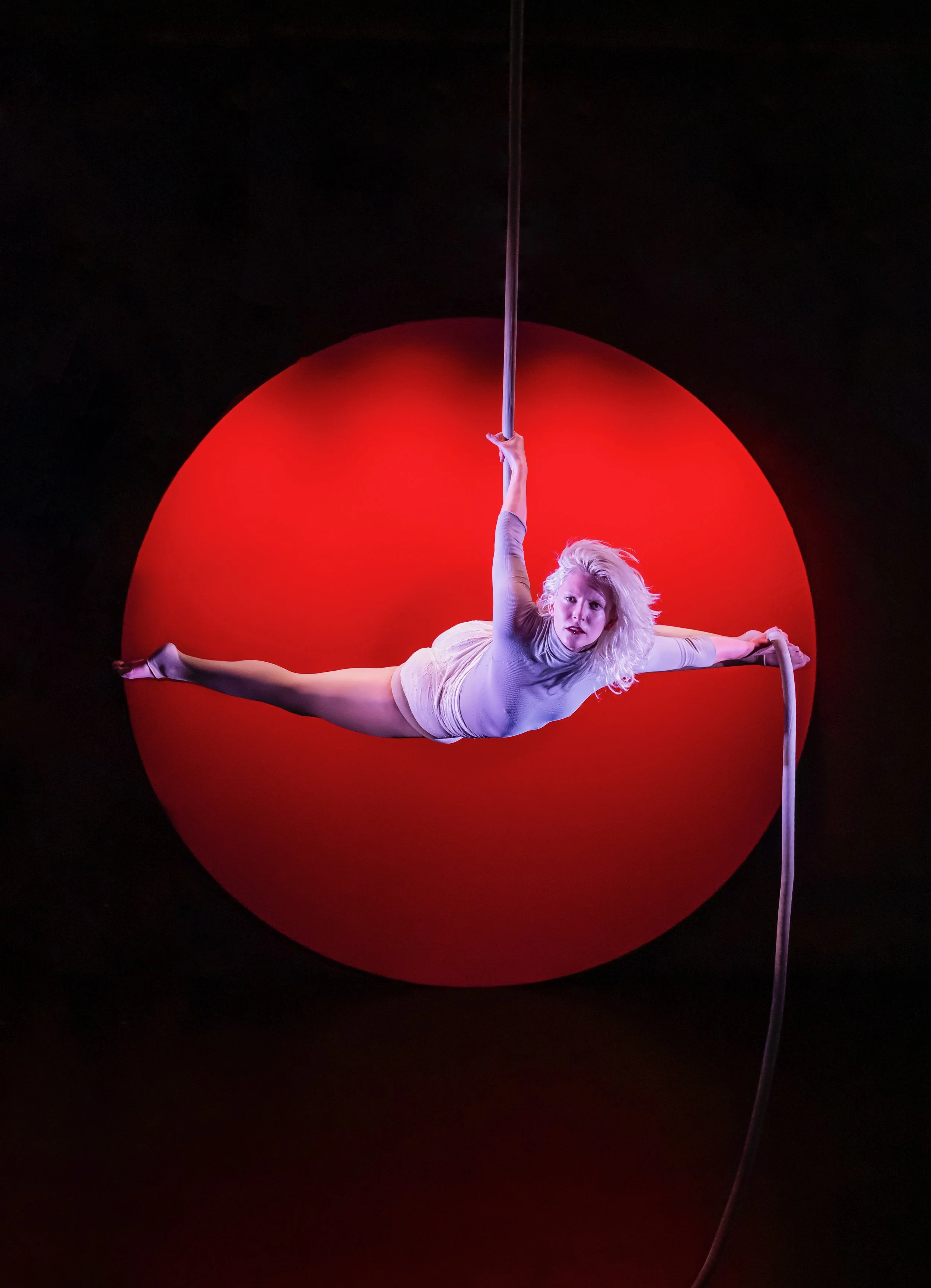 Aerial performer in a grey costume hanging horizontally from aerial silks against a red circular background.