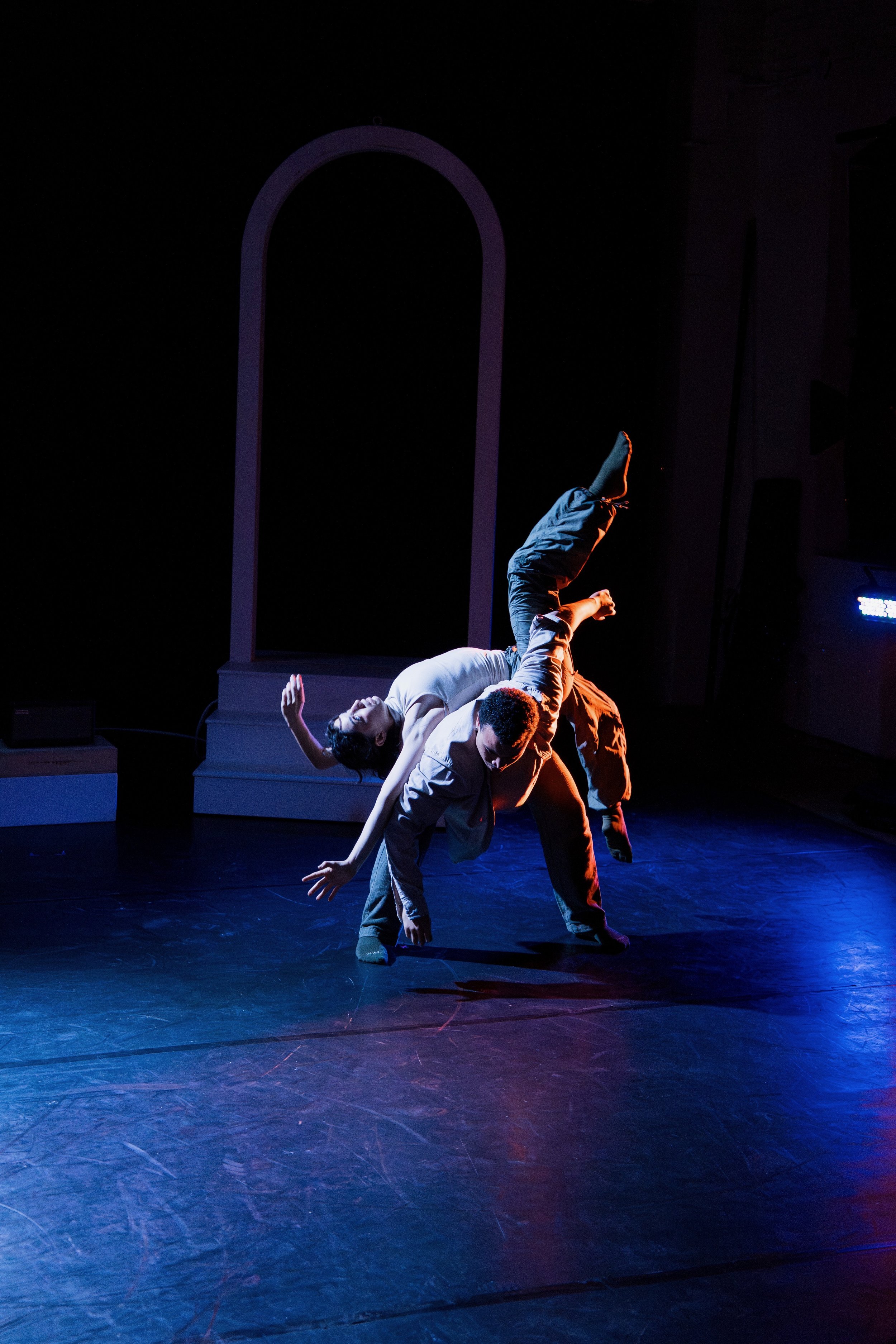 Two performers in a dance pose on stage, illuminated by colored lights, with a plain dark background and a decorative frame.