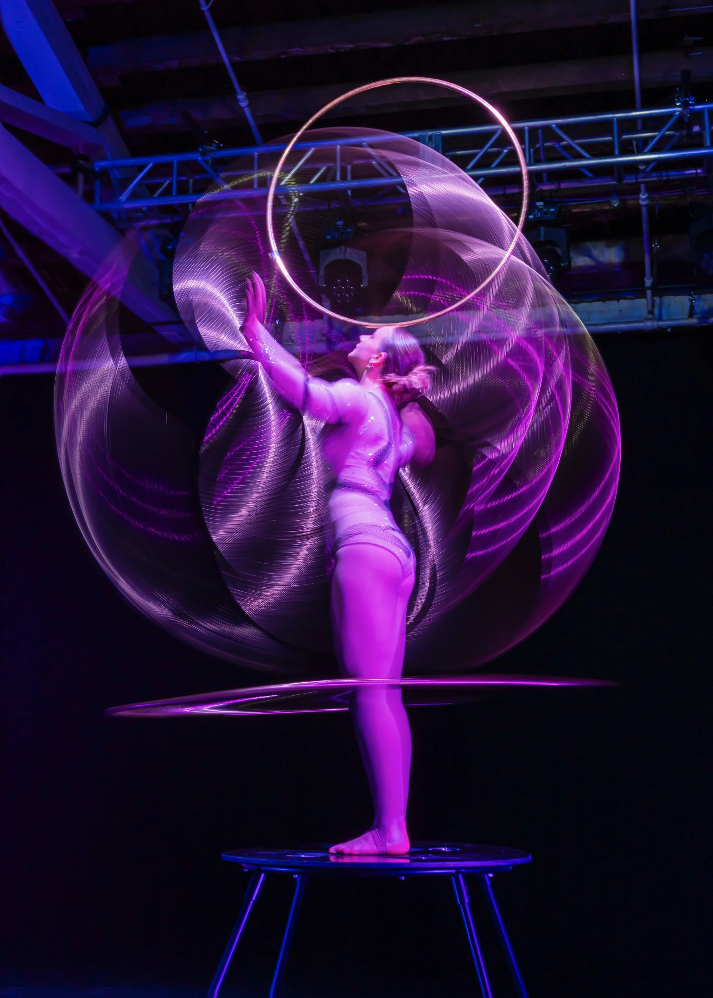 A performer on a small trampoline creating light trails with LED hoops, illuminated with purple and pink lights, in a dark setting.