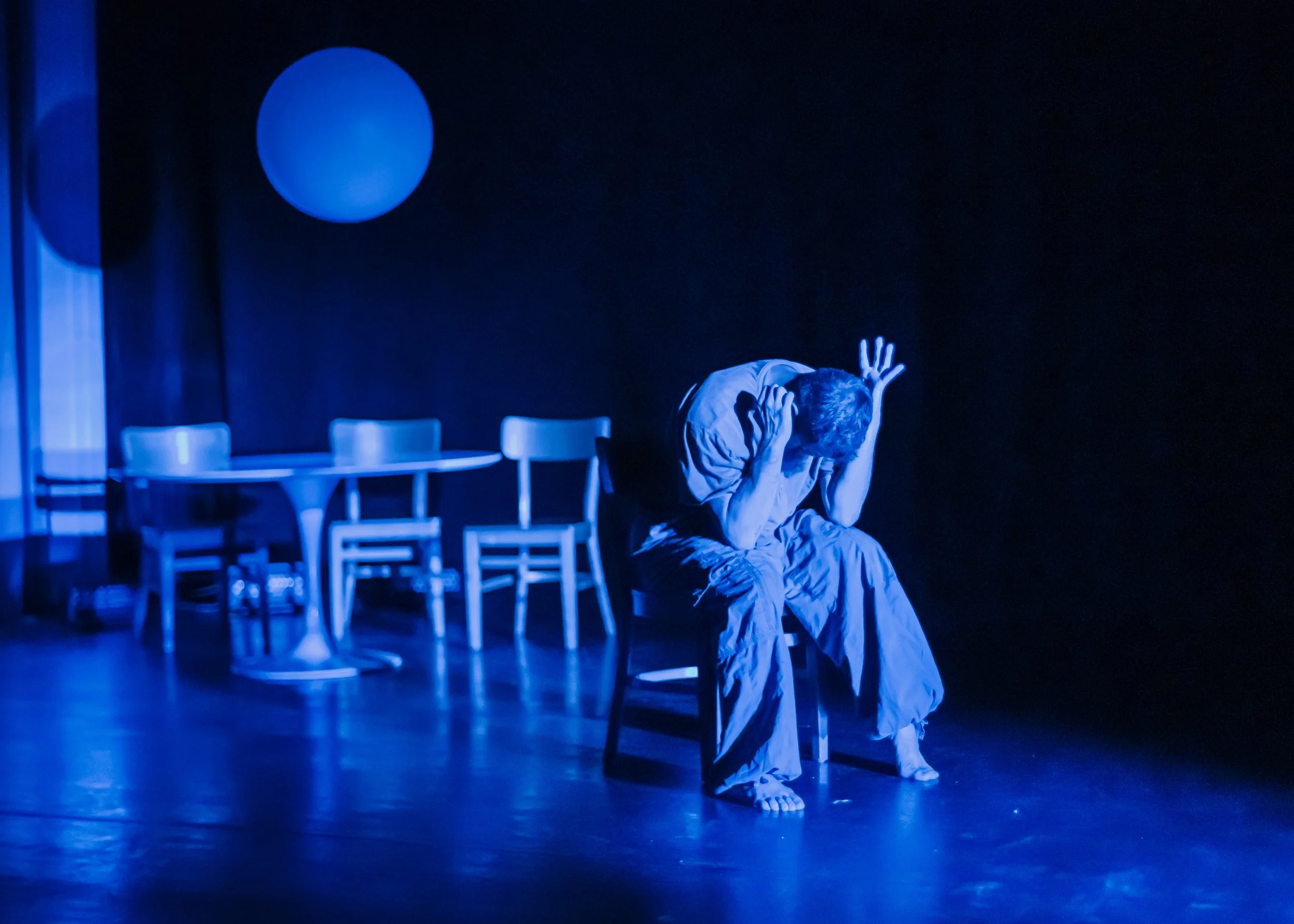 A person sitting on a chair with their head down and hands covering their face, illuminated by blue lighting on a stage with a table, four chairs, and a large round light in the background.