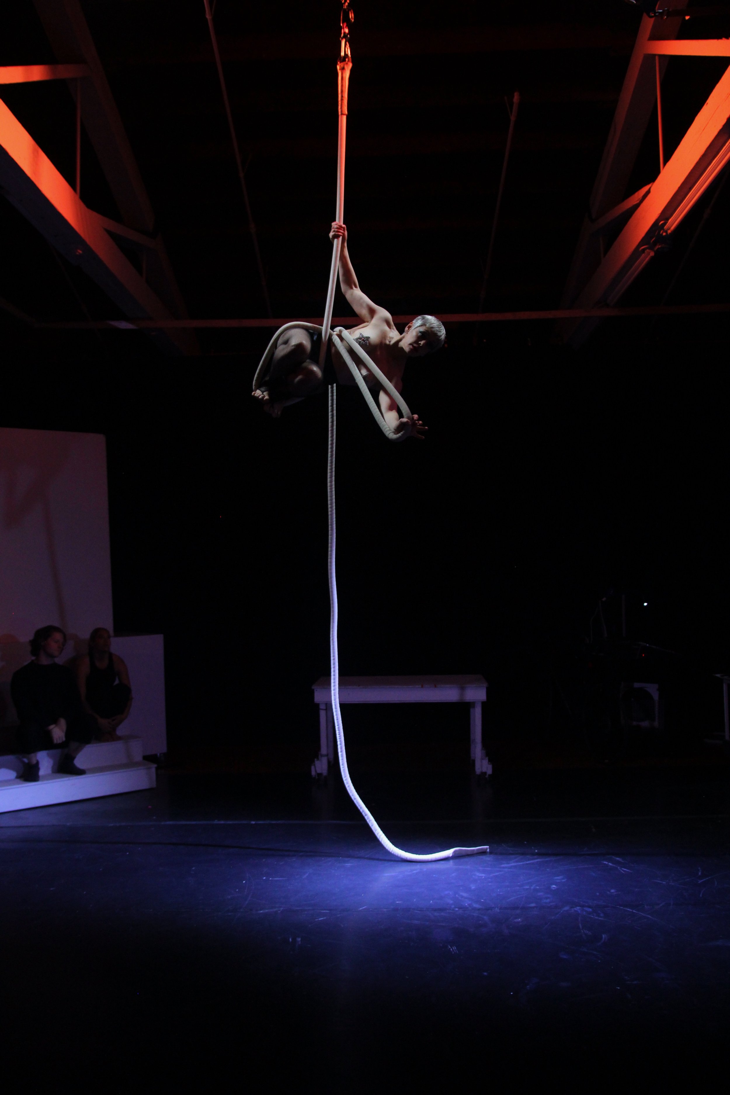 A performer is hanging upside down on a pole, holding a rope with one hand, during a circus or acrobatic show, with two spectators seated in the background.