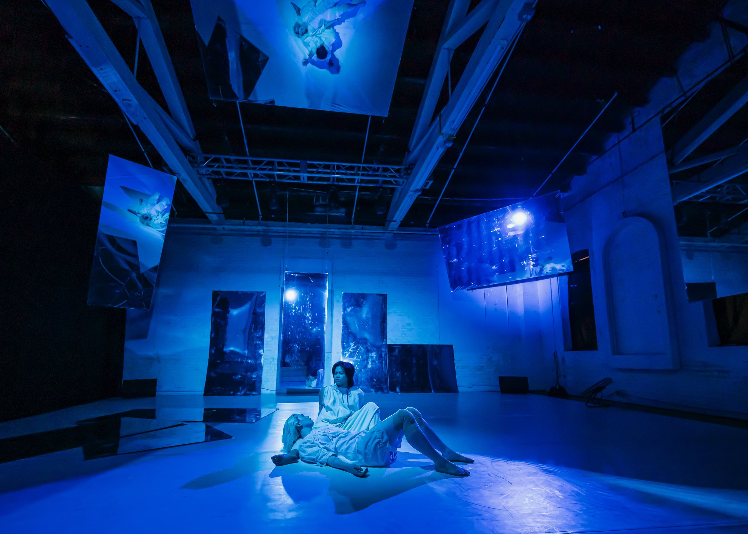 Two women are in a dimly lit room illuminated by blue light, one lying on the floor and the other sitting beside her, both dressed in white. The room features hanging and leaning reflective panels and banners that show similar abstract reflections, c