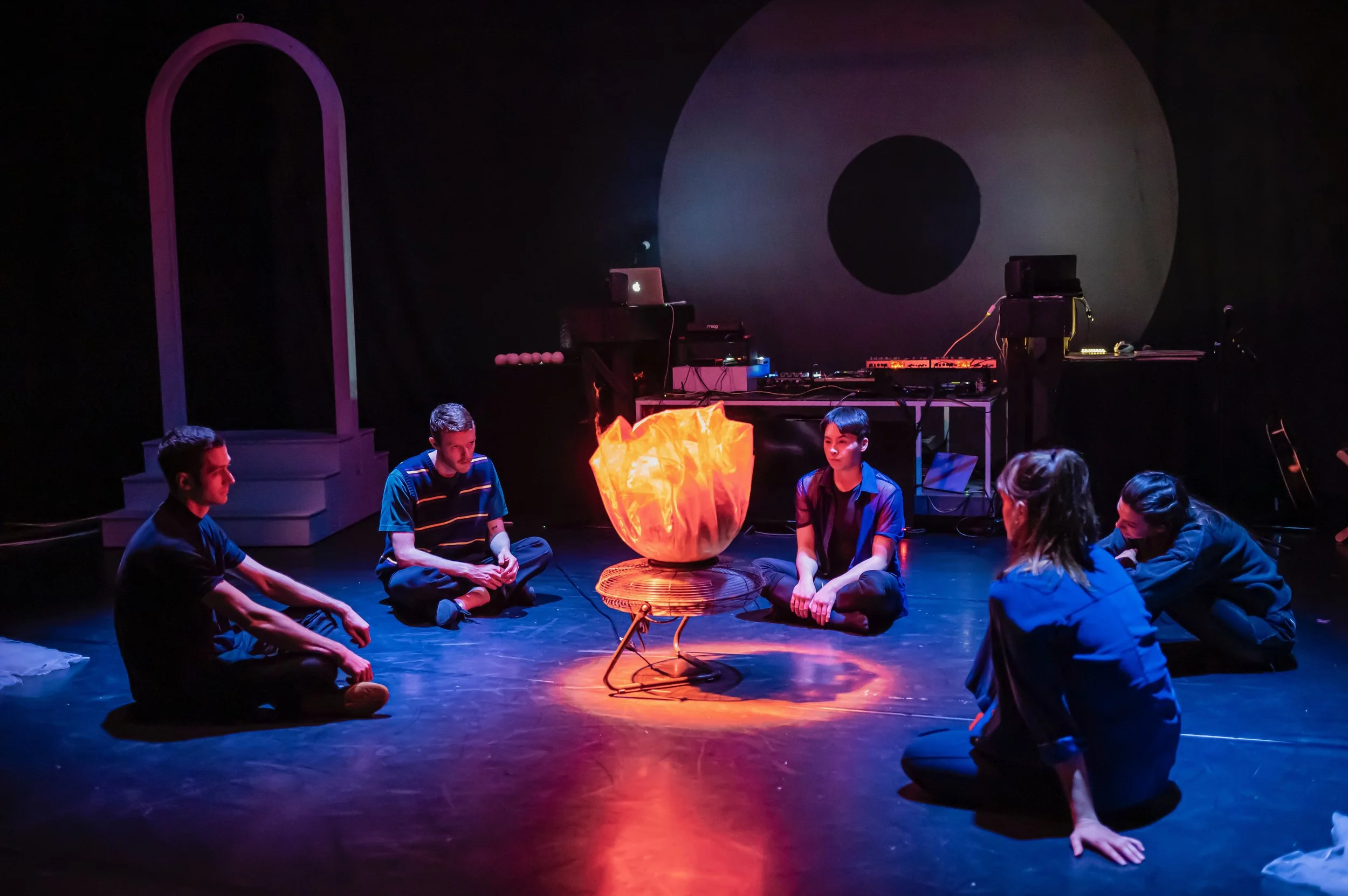 Five people sitting cross-legged in a circle around a glowing, fiery bowl on a stand in a dark room with artistic lighting, with DJ equipment and a large circular backdrop in the background.