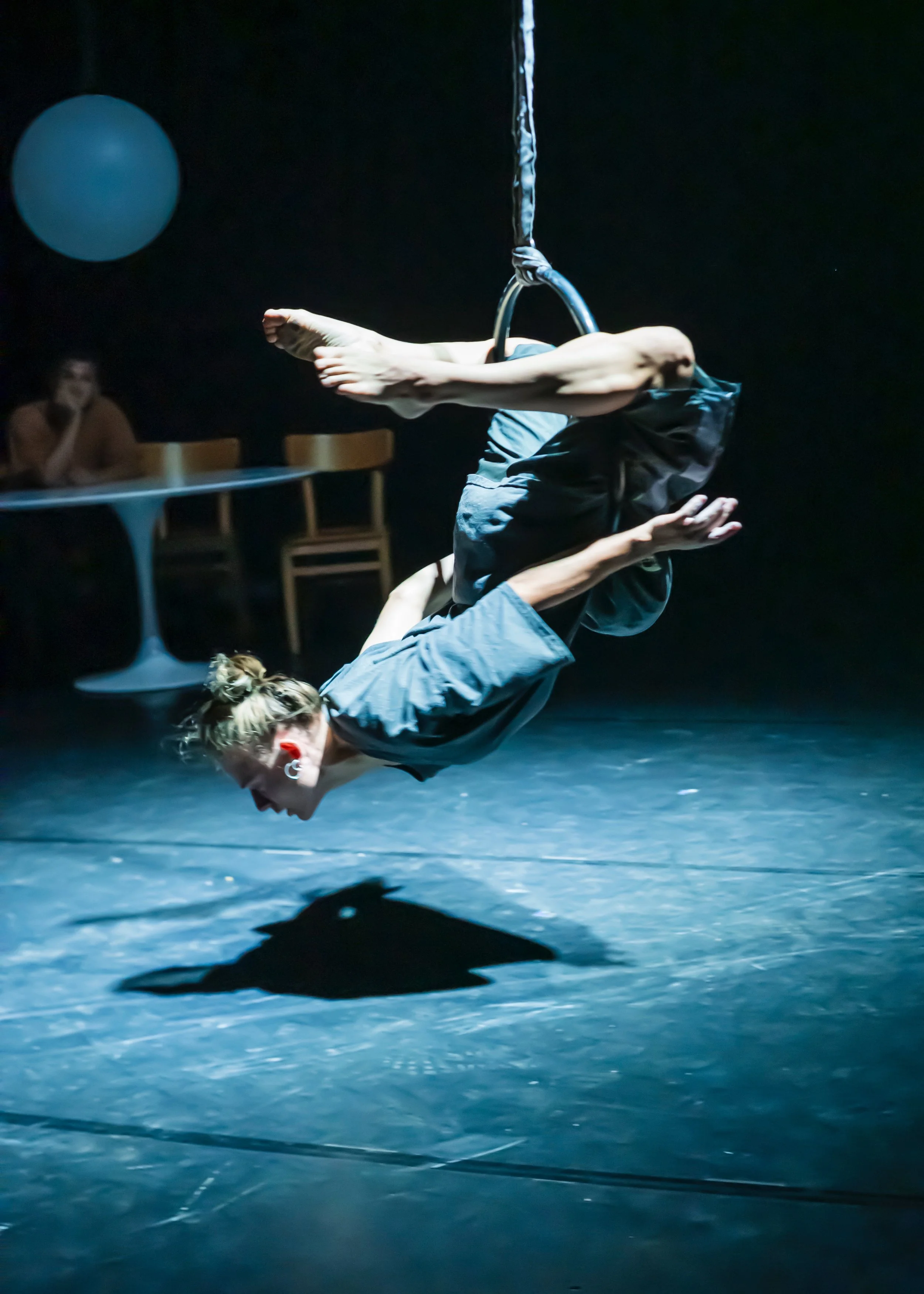 A female performer in mid-air on an aerial hoop, performing an acrobatic routine on a dark stage with a spotlight, with a person watching in the background.