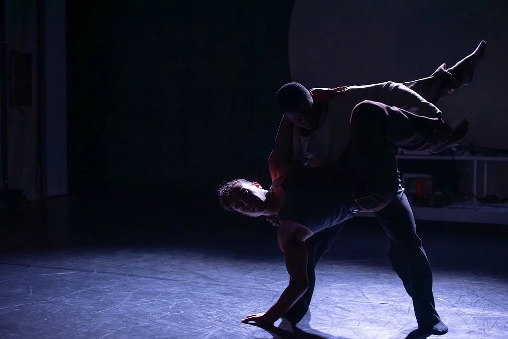 Two male performers practicing acro yoga or a similar physical routine on a dimly lit stage, with one lifted and supported by the other.