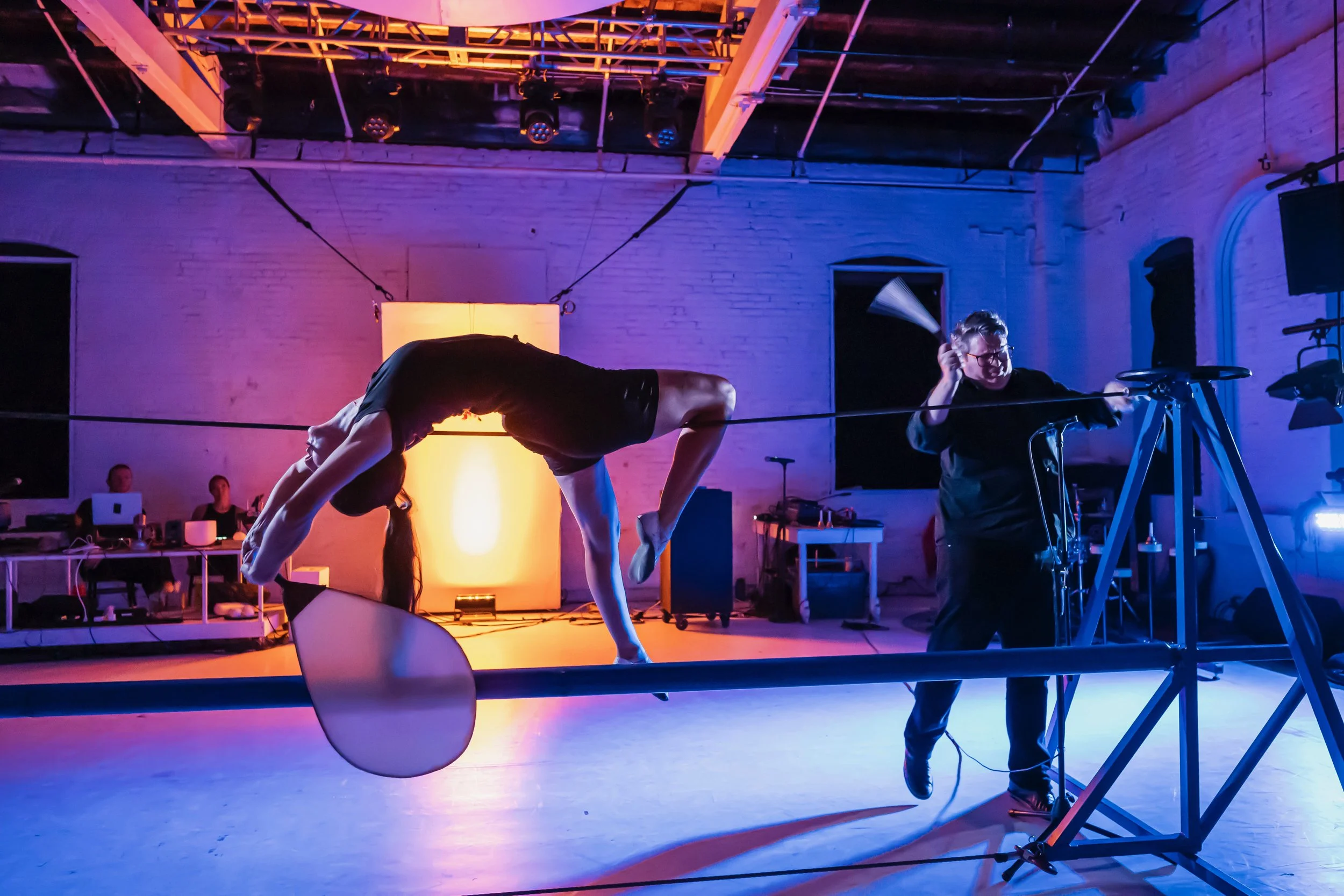 A woman performing a contortion pose on a slackline setup inside a room with colorful lighting; a man operating a camera on a tripod nearby; people working at computers in the background.