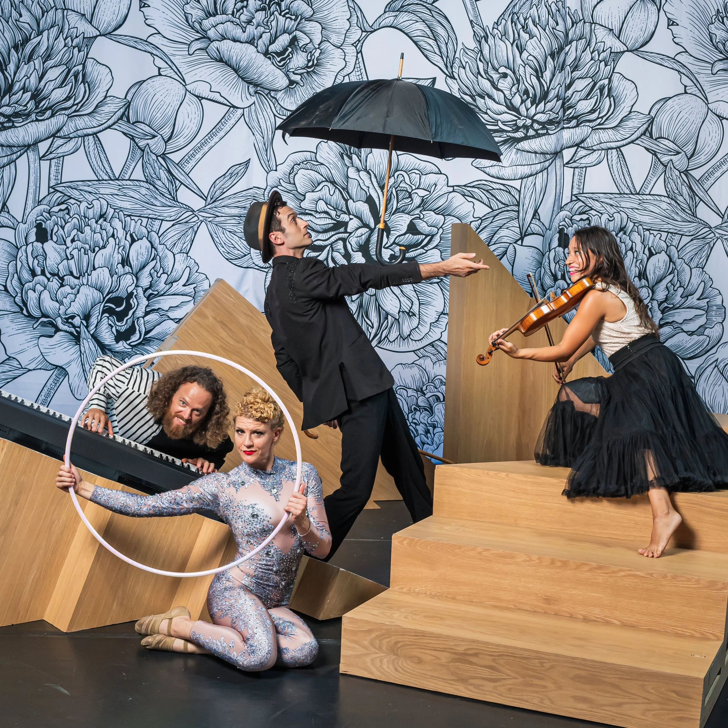 Four performers on stage: a woman playing a violin and a man reaching toward her, one person kneeling holding a hoop and a keyboardist leaning into the hoop, in front of a floral mural.