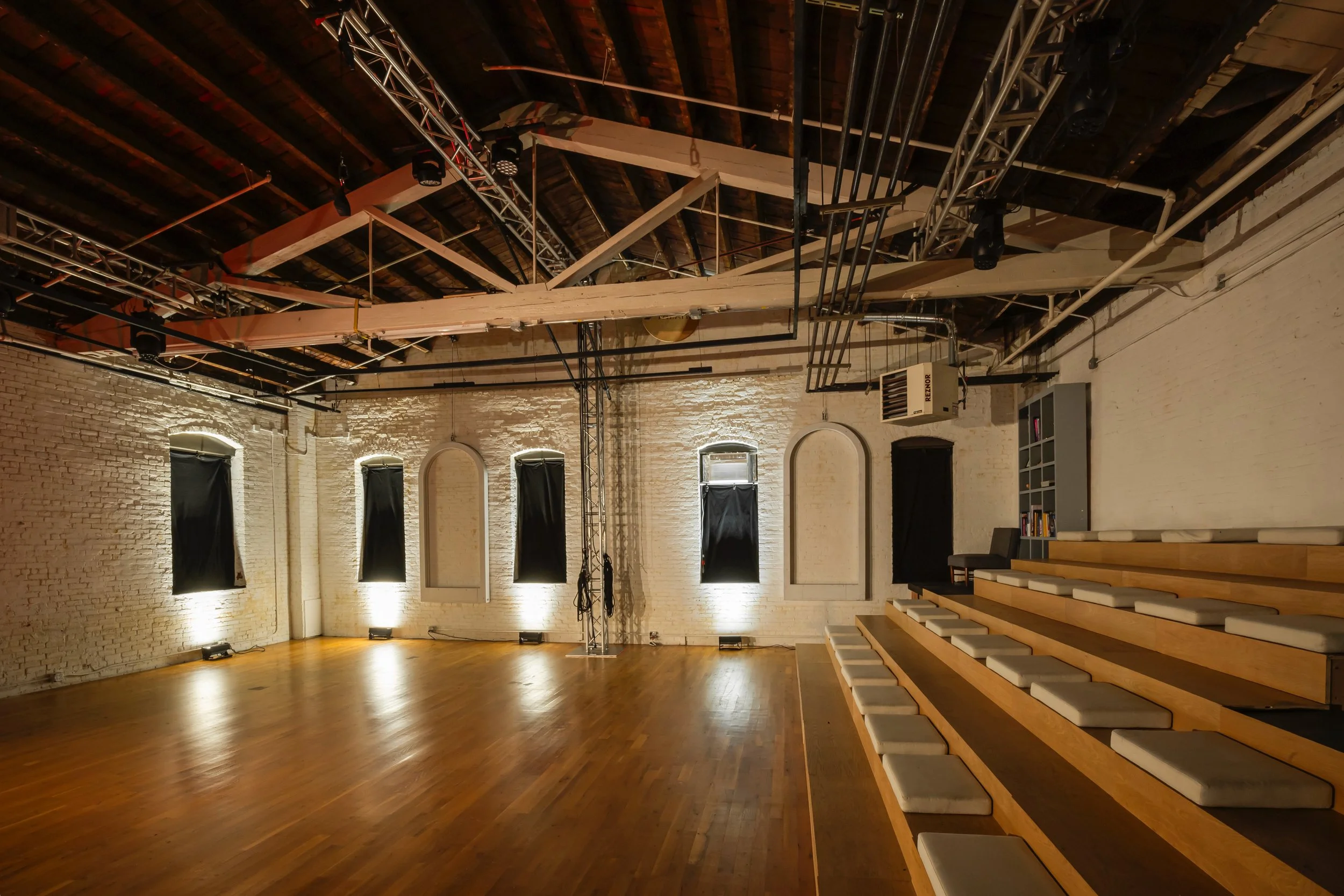 Empty performance or event space with wooden floor, white brick walls, black curtains on windows, stage lighting, and tiered seating with cushions to the right.