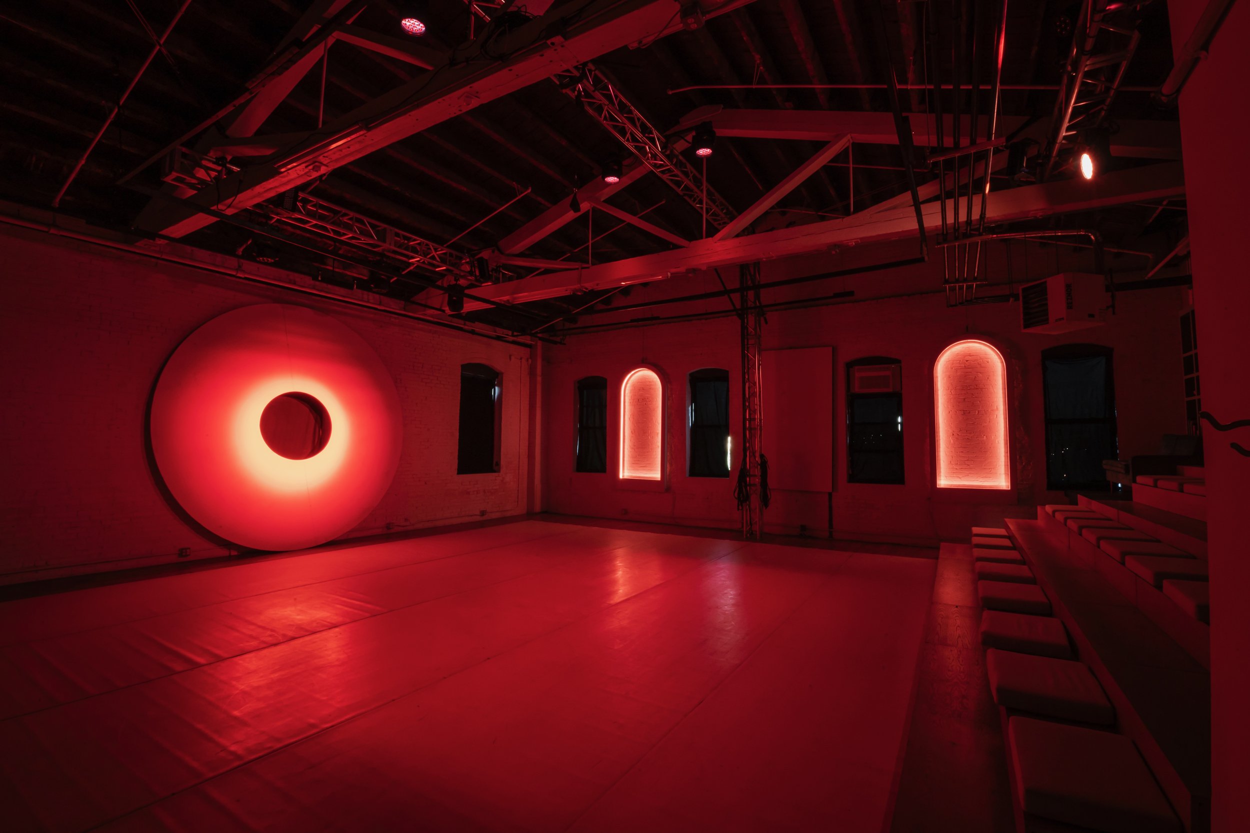 A dimly lit room with red lighting, featuring a large circular art installation on the wall and several arched windows illuminated with red neon lights. There are chairs along one wall and exposed ceiling beams and structures above.