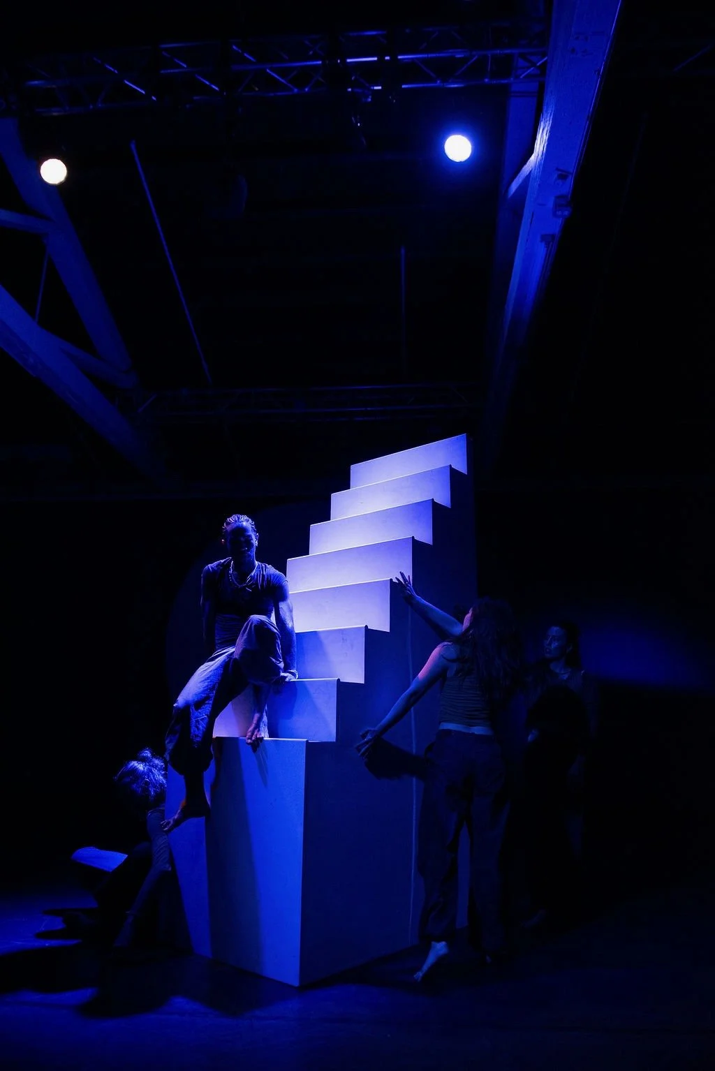 Group of performers on stage with a staircase prop, illuminated in blue light, during a theatrical performance.