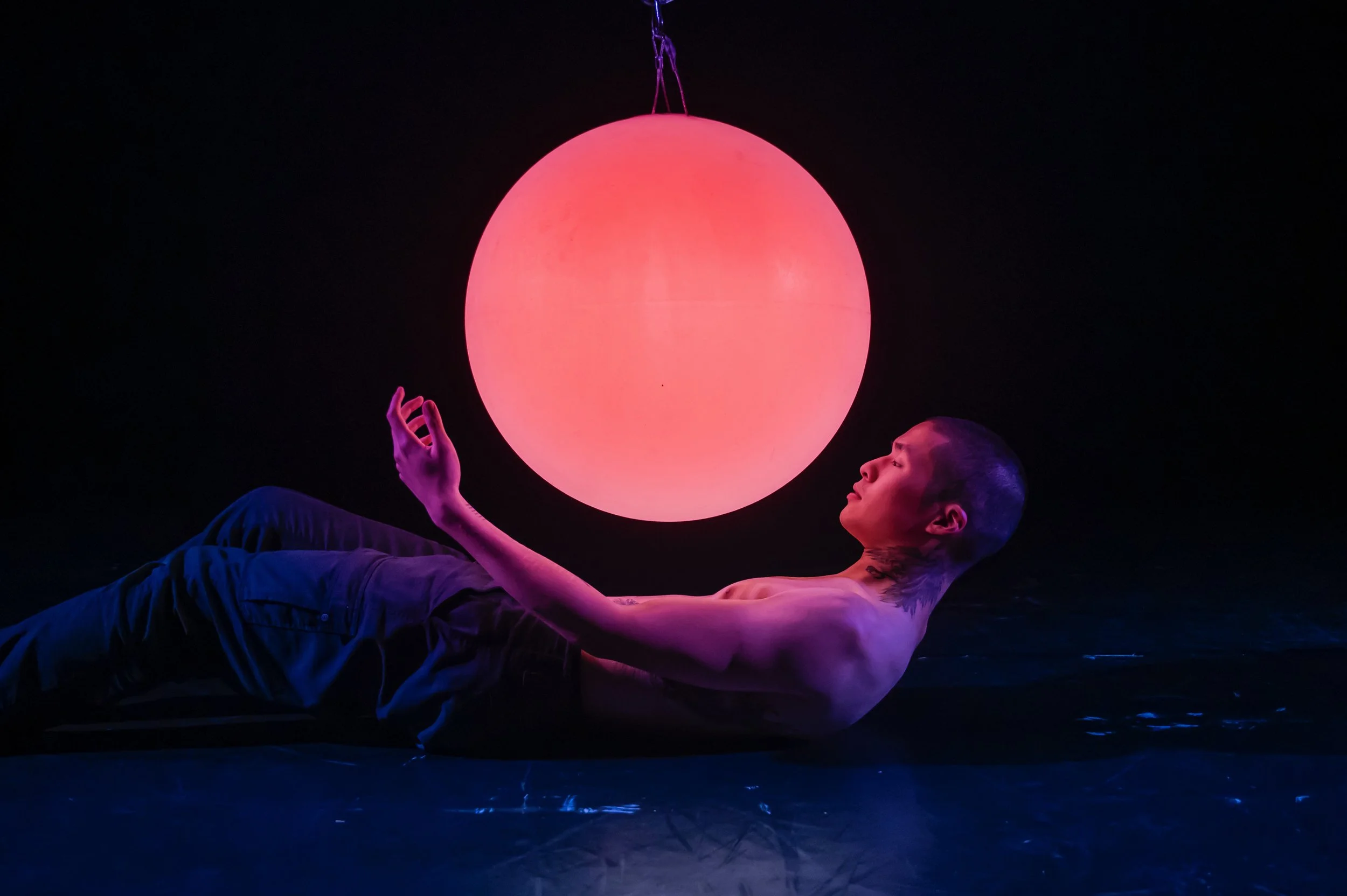 A shirtless person with short hair and tattoos lying on a dark floor, looking upward at a large glowing pink and purple sphere hanging above them.