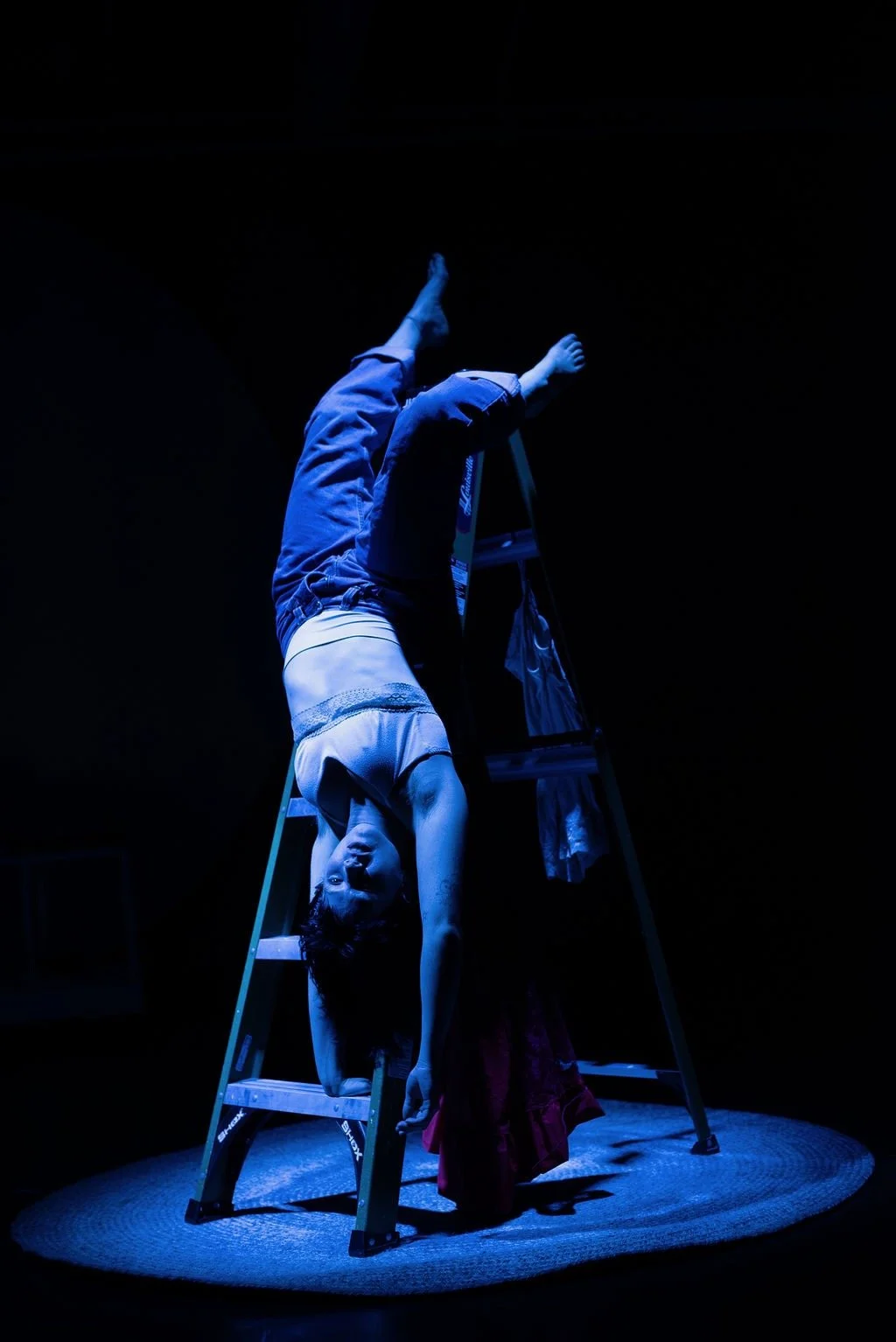 Person performing a handstand on a ladder in a dark room, illuminated by blue light.