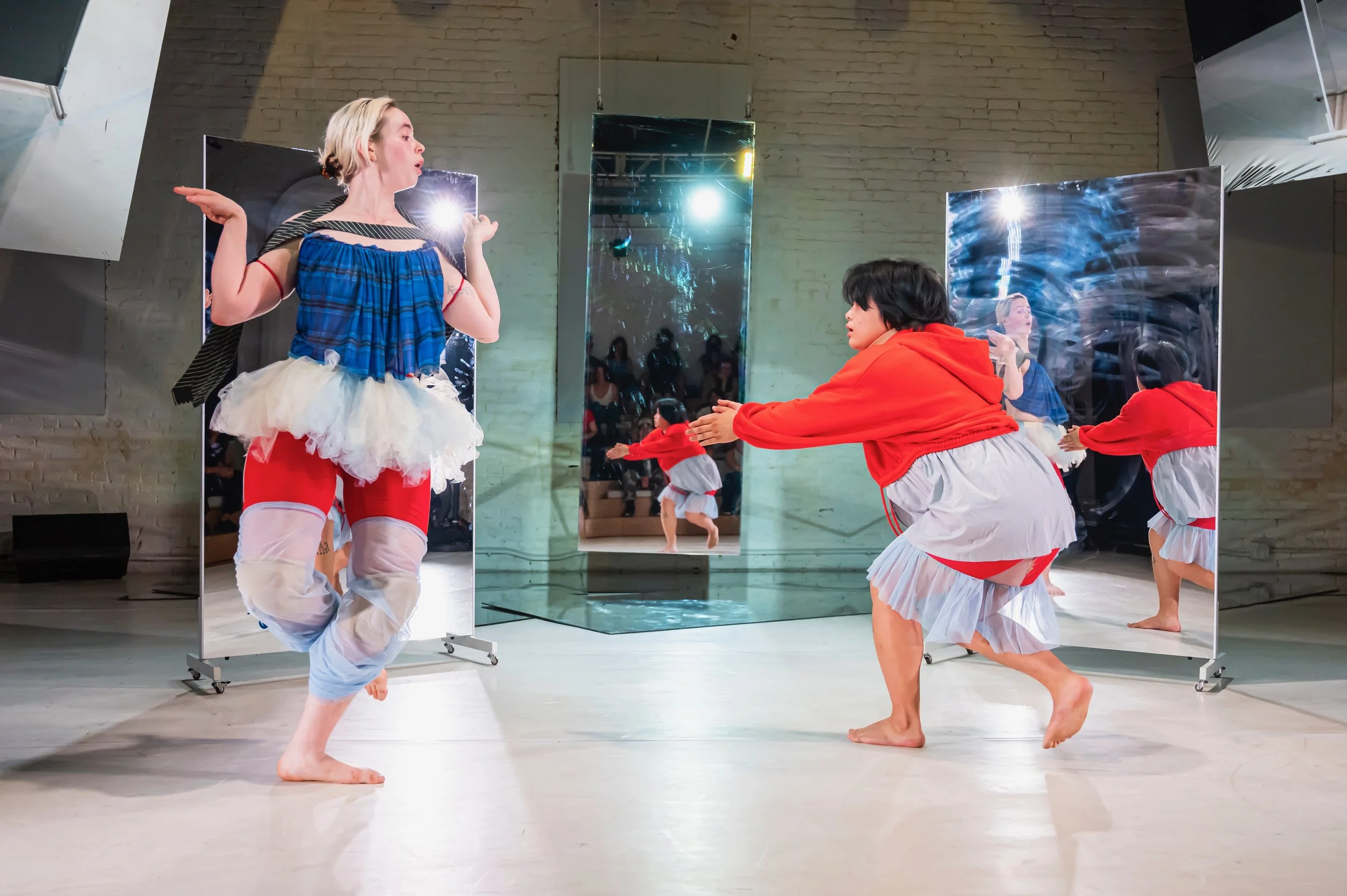 Two young women dancing in front of mirrors in a dance studio. One woman is wearing a blue top, a tutu, and ripped pants, while the other woman is dressed in a red hoodie and ruffled skirt.