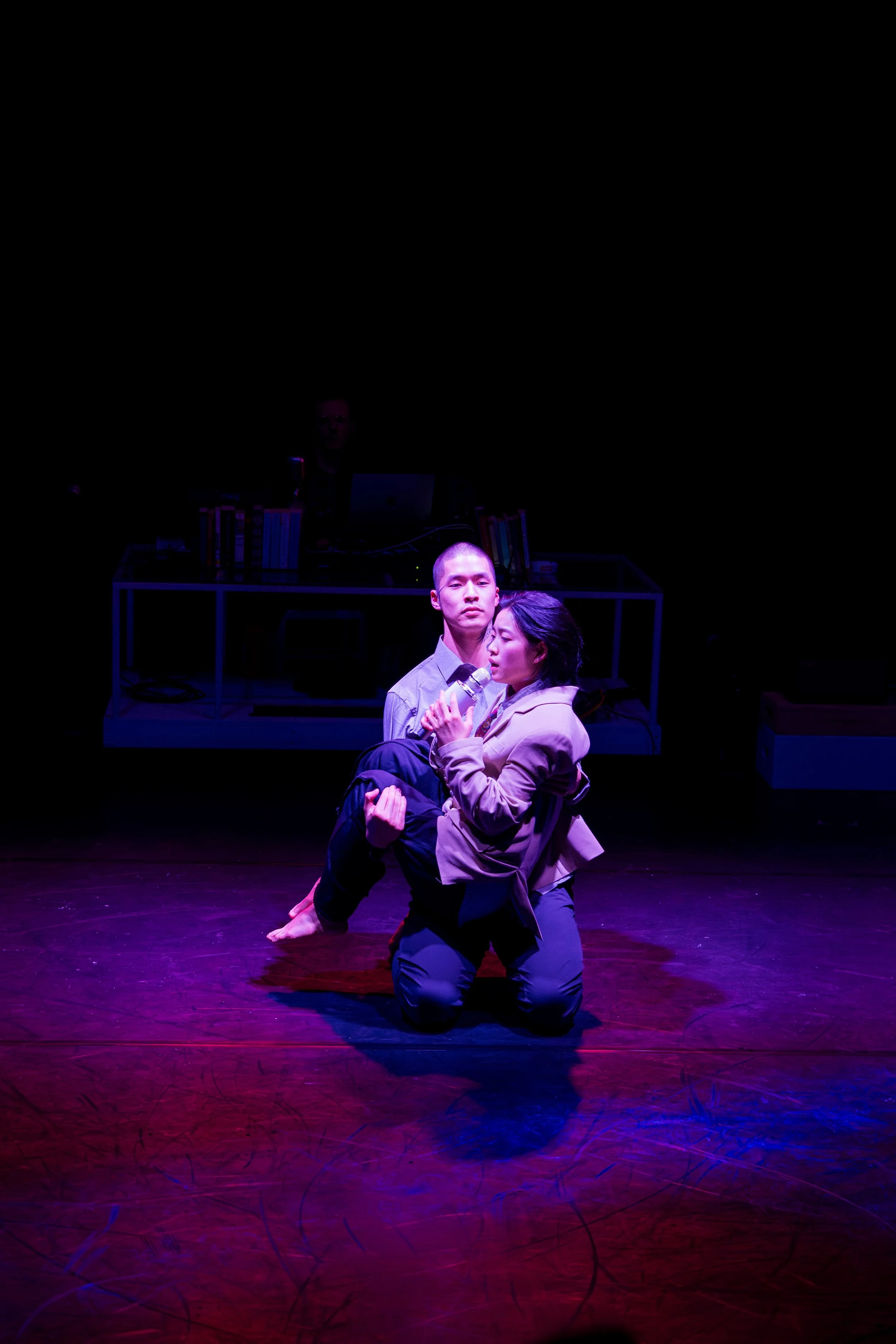 Two performers kneel on a stage under purple lighting, with one holding a microphone and the other with fists clenched, while a DJ and equipment are visible in the background.