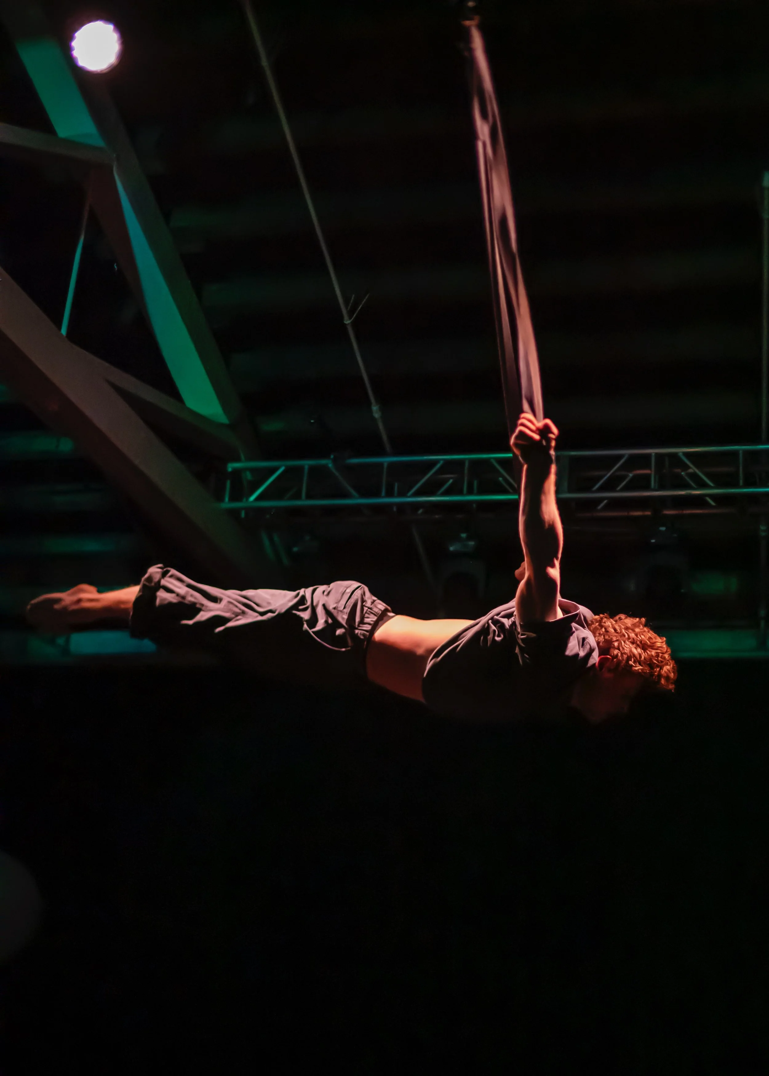 A person with curly hair is performing an aerial silk act, hanging horizontally in a dark setting with stage lights, holding onto a long fabric